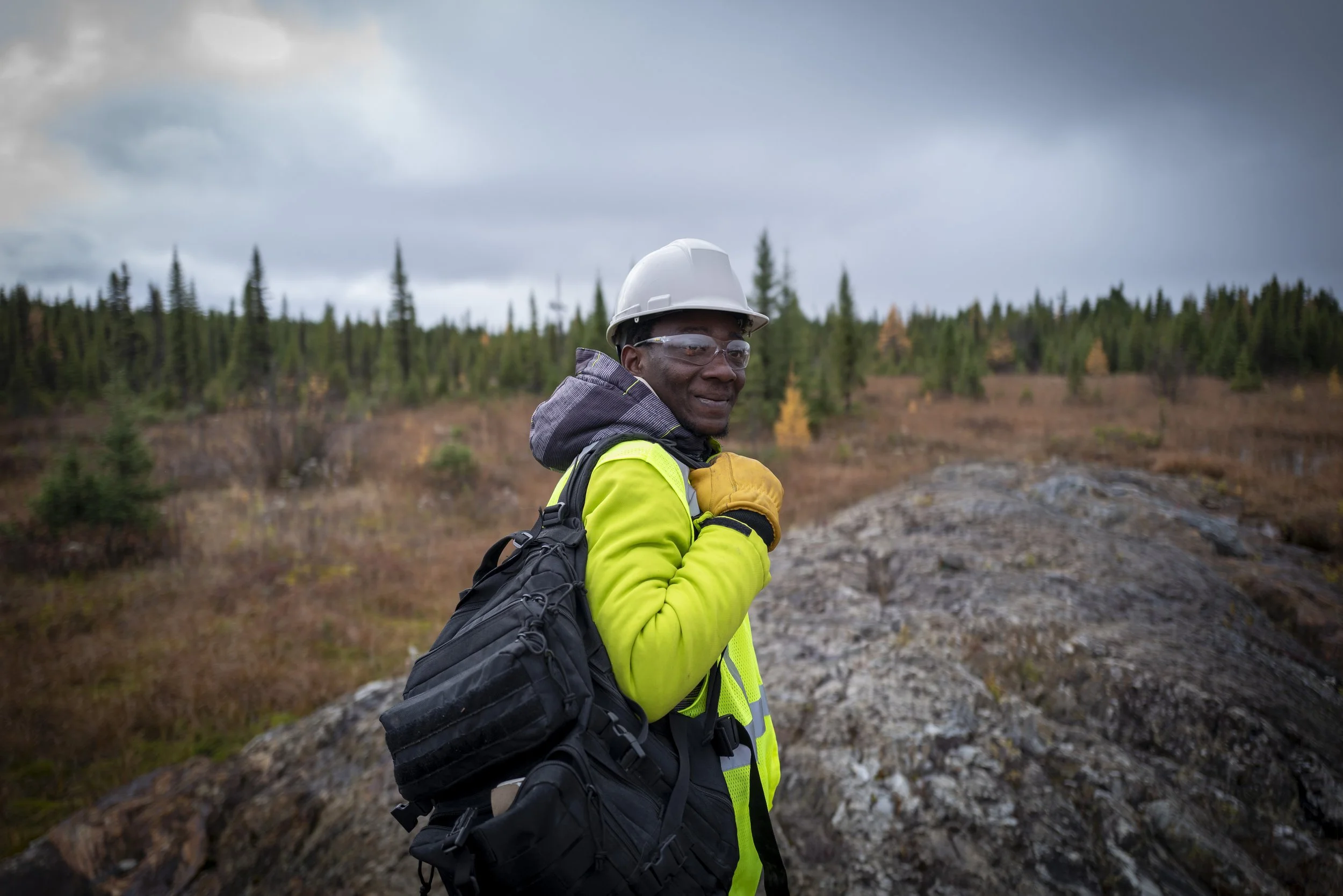 A man in a high-visibility yellow jacket and white safety helmet stands outdoors with a backpack, in a forested area under a cloudy sky.