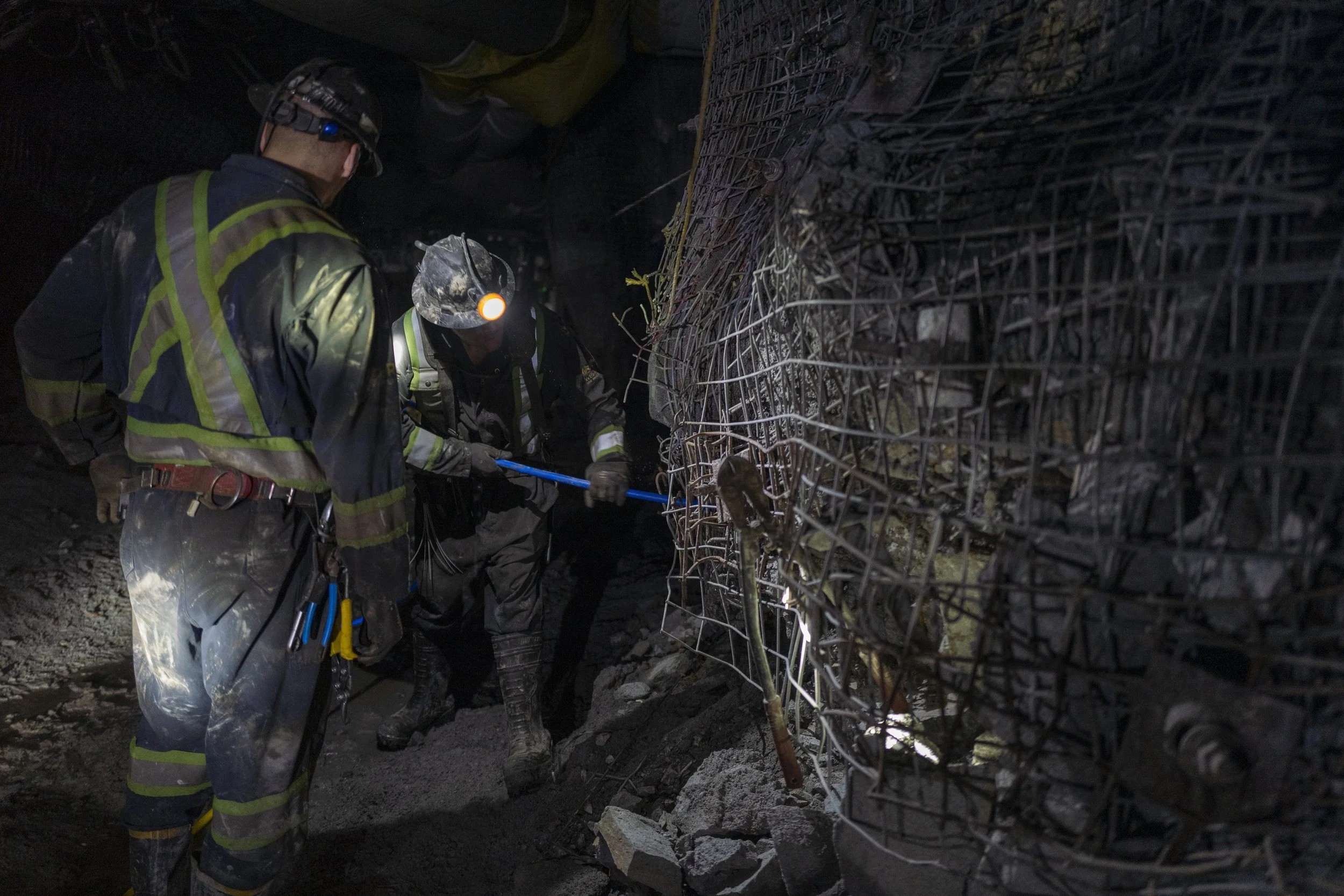 Two construction workers in safety gear working inside a tunnel reinforced with wire mesh and rebar, illuminated by headlamps and flashlights.