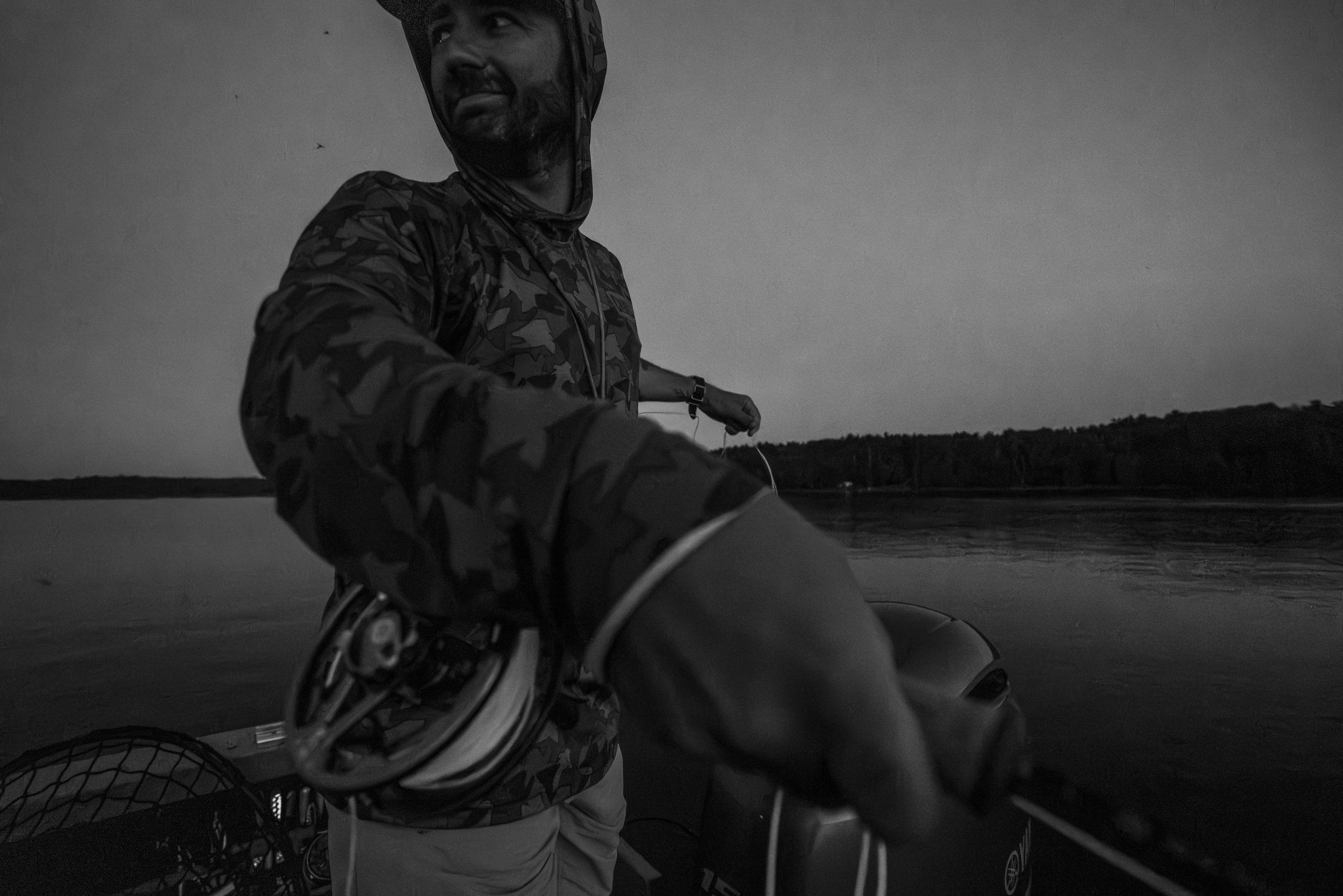 A man in camouflage clothing on a boat, holding a fishing rod, smiling and looking away, with a body of water and a tree-lined shoreline in the background during dusk or dawn.