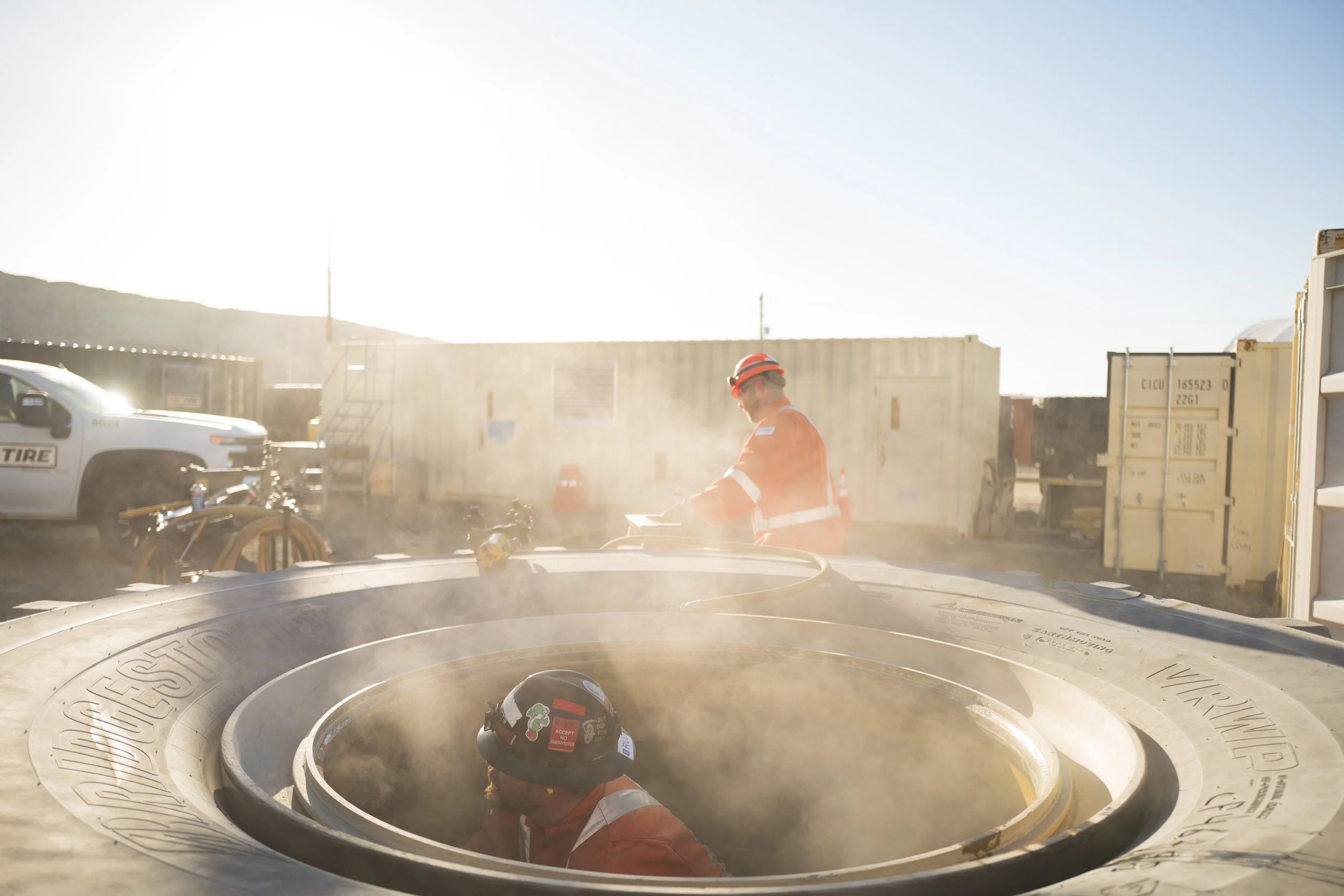 Two workers in orange safety gear and helmets working on a large industrial tire outdoors, with one worker partially inside the tire and the other standing nearby, glowing sun and containers in the background.
