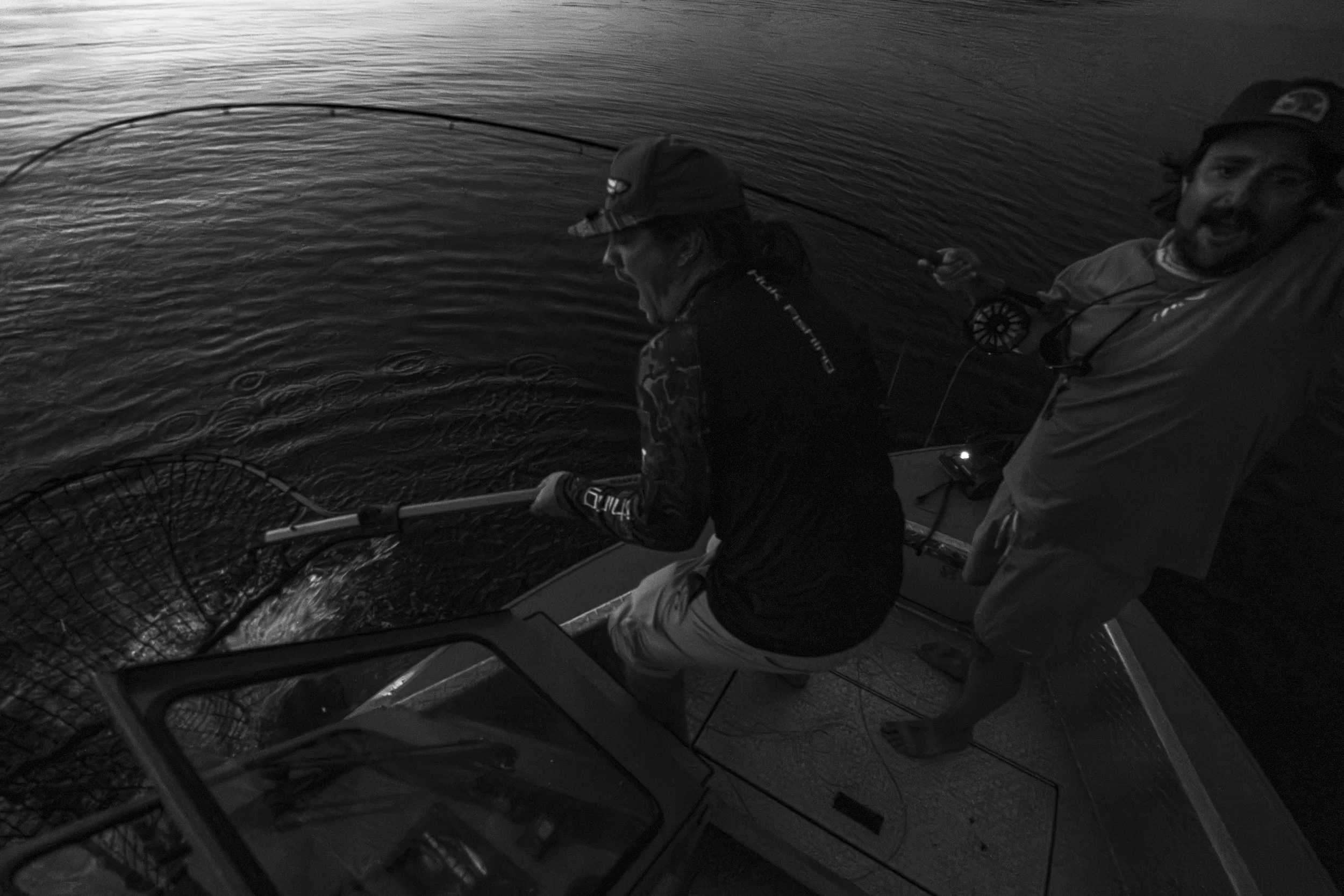 Two people fishing in a boat on a body of water, one is drawing in a fishing net, both are wearing hats and outdoor clothing, nighttime setting.