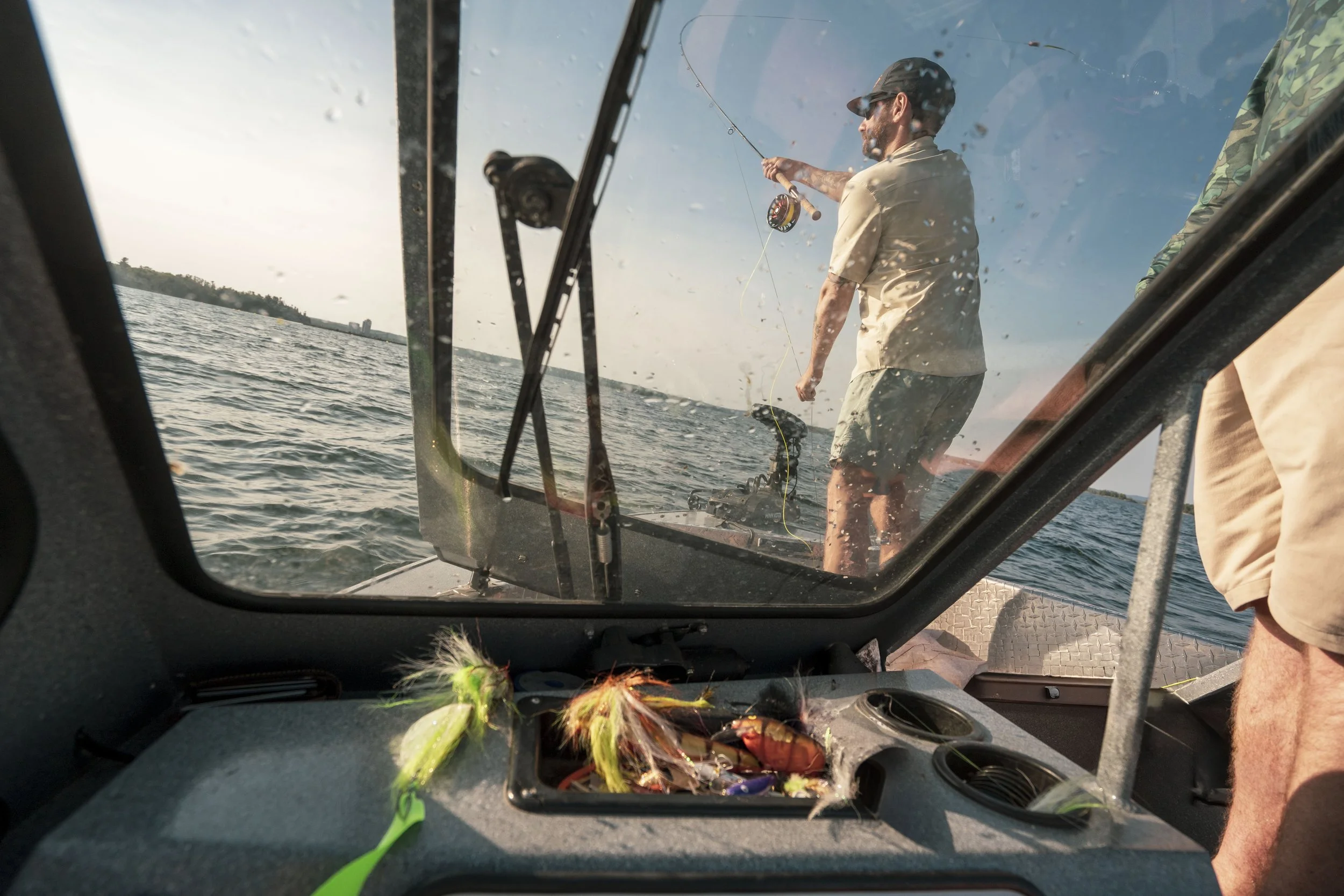 View from inside a boat showing a man fishing on the water, seen through a windshield with fishing gear and tackle in the foreground.