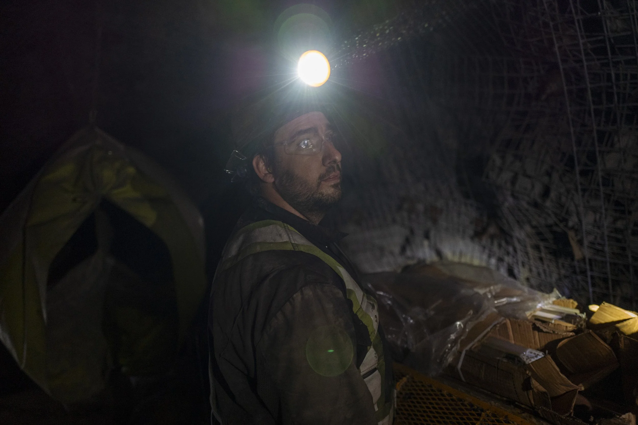A male worker wearing a headlamp inside a tunnel construction site, surrounded by metal scaffolding and construction materials.
