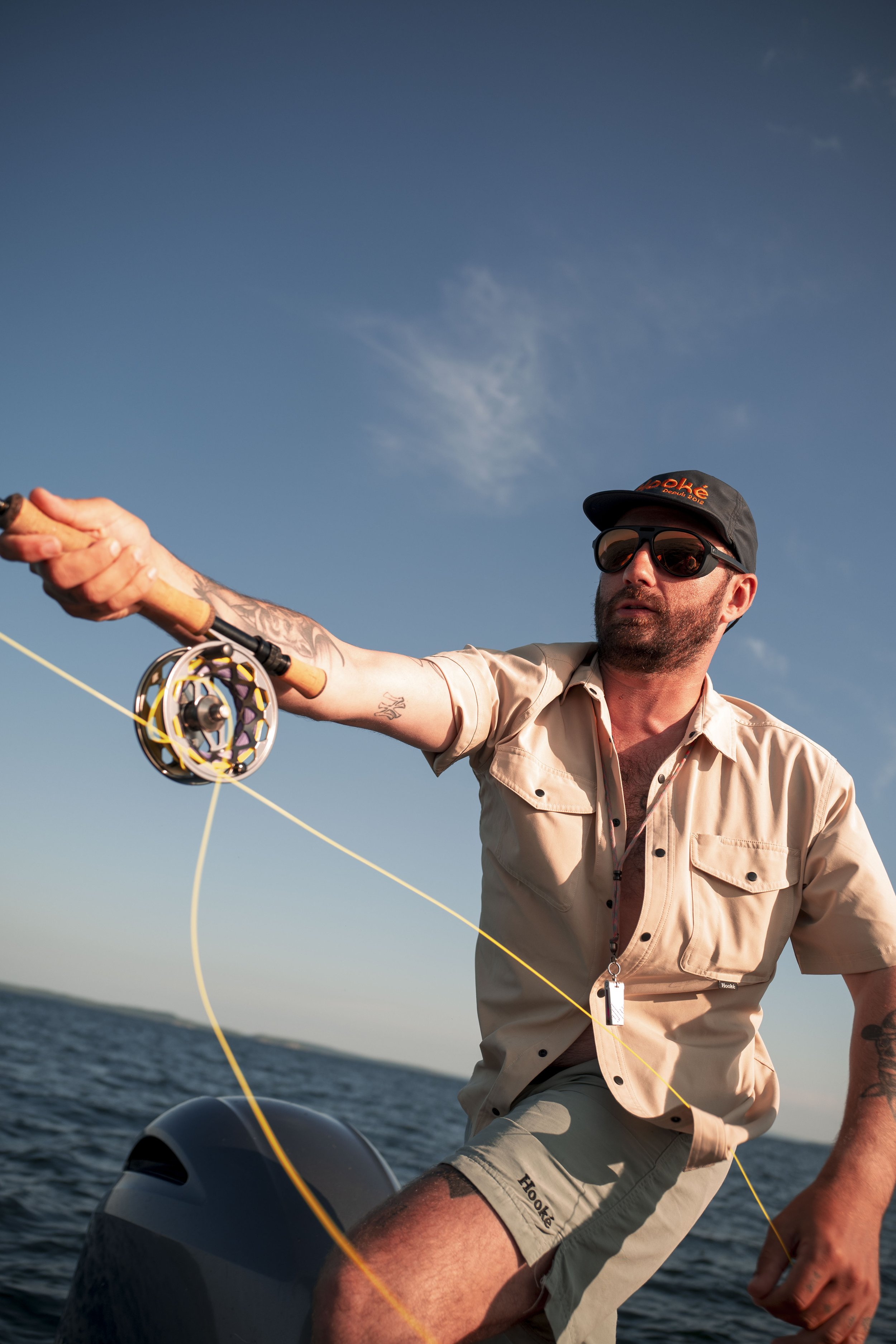 A man fishing on a boat, wearing sunglasses, a baseball cap, a beige shirt, and khaki shorts, holding a fishing rod with a reel, with water and sky in the background.