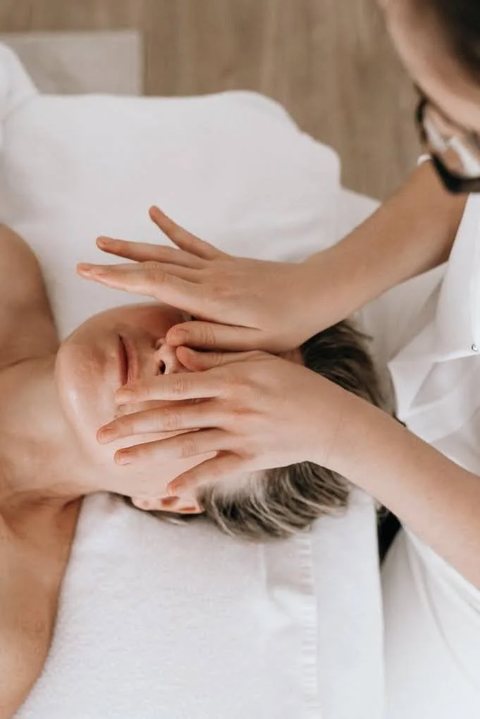 Une personne reçoit un soin du visage ou un massage facial, avec un professionnel appliquant une pression douce sur le visage dans un environnement de spa ou de clinique.