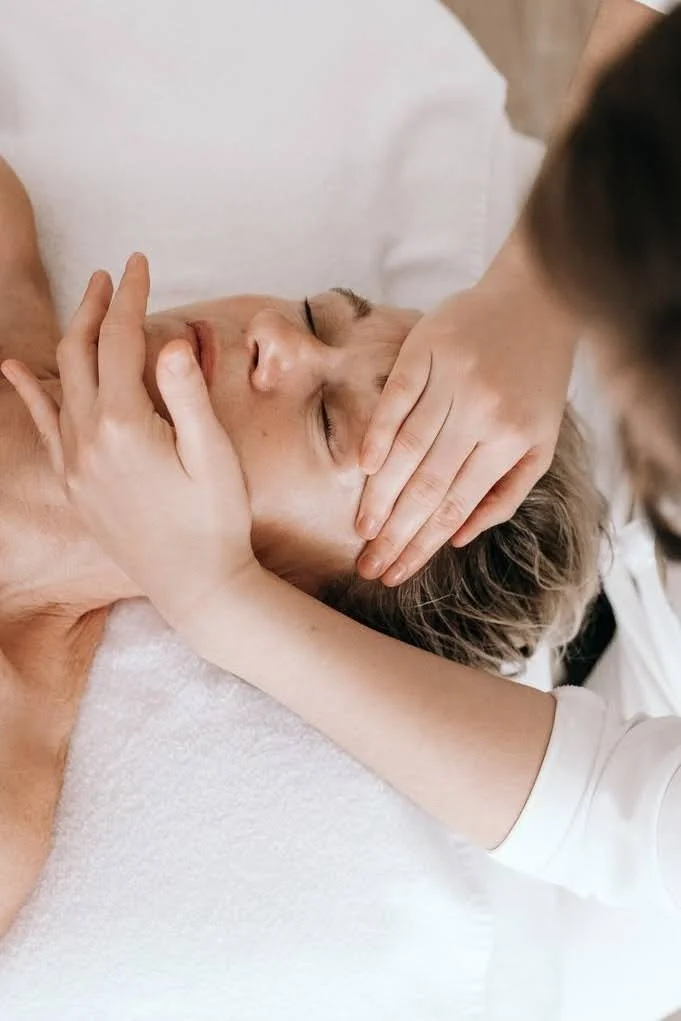 Une personne reçoit un massage facial, allongée sur une table avec un drap blanc, les yeux fermés, tandis qu'une autre personne lui masse le visage.