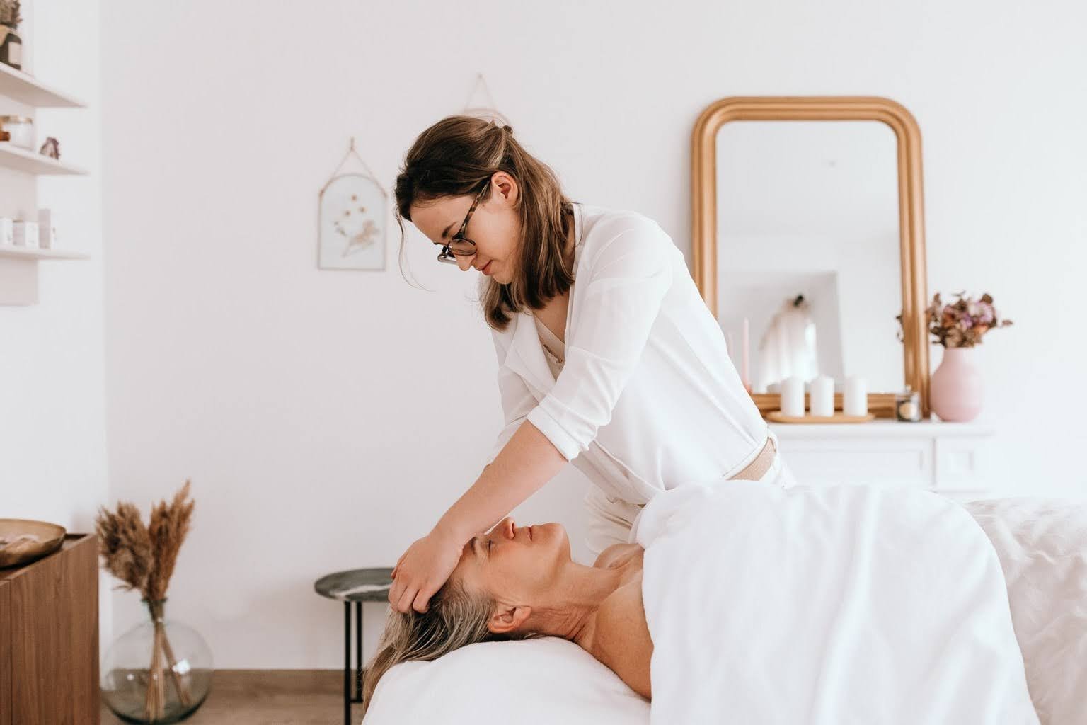 Une femme donnant un massage de tête à une autre femme allongée dans un lit, dans une chambre décorée avec un miroir, des bougies et un vase de fleurs.