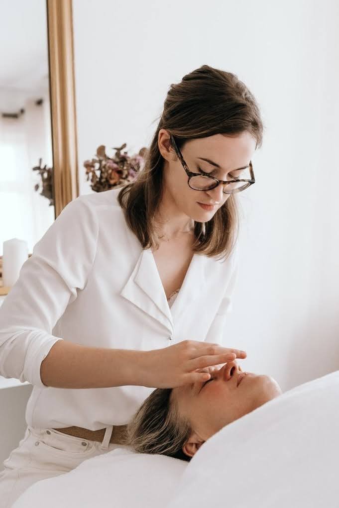Une femme procure un soin de spa ou de massage facial à une autre femme allongée sur un lit. La femme qui donne le soin porte des lunettes et une chemise blanche, tandis que la personne recevant le soin est allongée avec les yeux fermés.