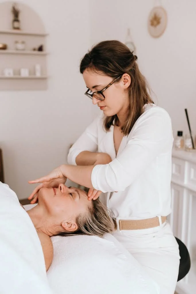 Une esthéticienne ou une thérapeute effectue un soin facial à une femme âgée allongée, dans une pièce sobre et élégante.