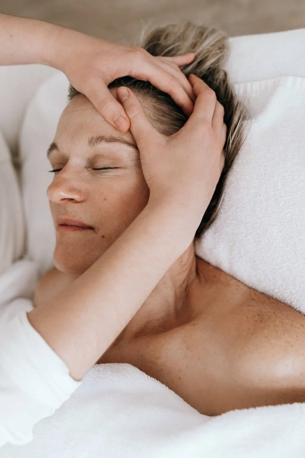 Une personne reçoit un massage dans un lit blanc, axée sur ses épaules et sa tête, avec un visage détendu et les yeux fermés.