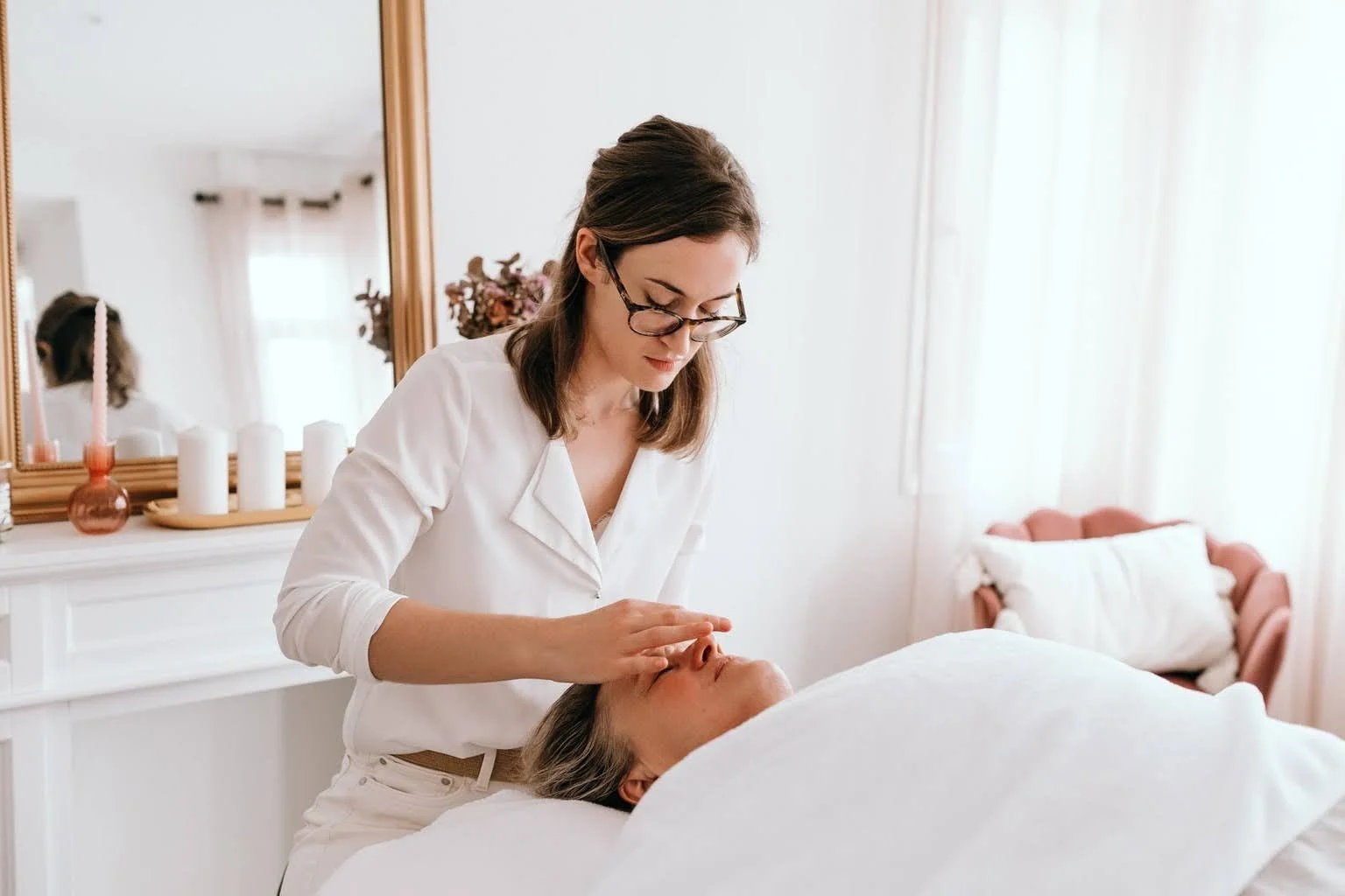 Une esthéticienne pratique un soin du visage à une cliente allongée sur une table de massage dans une pièce lumineuse.