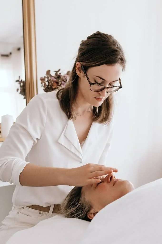 Une femme donnant un soin du visage à une autre femme allongée, dans un environnement calme. La femme qui donne le soin porte des lunettes et un vêtement blanc.