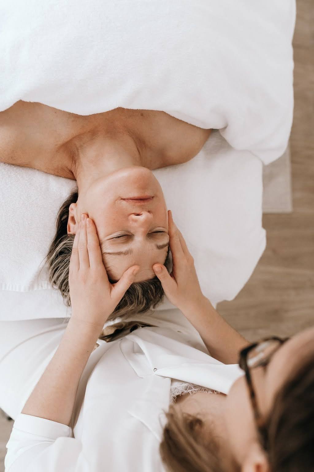 Une femme reçoit un massage de la tête dans une ambiance relaxante, allongée sur un lit blanc avec une couverture blanche.