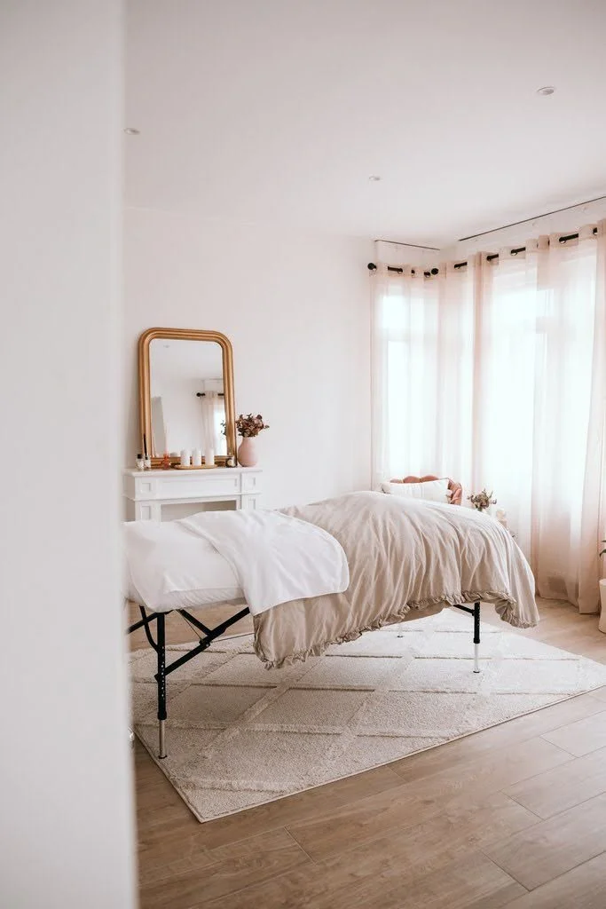 Une chambre avec un lit beige, des rideaux blancs, un miroir doré, et une petite table avec des décorations, éclairée par la lumière naturelle.