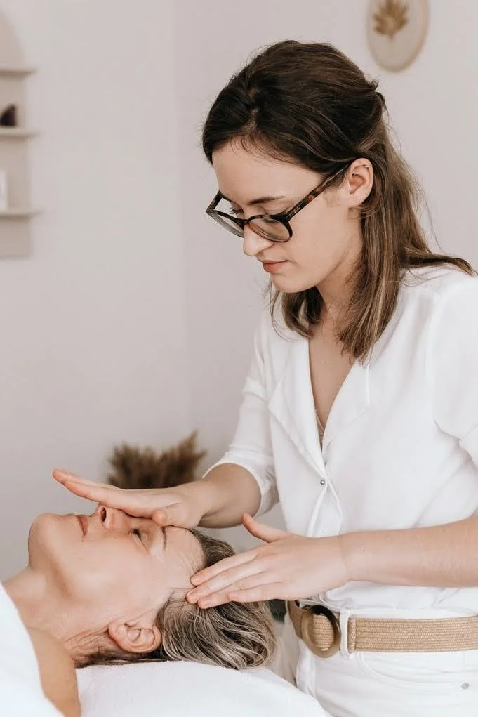 Une femme effectue un soin du visage ou une manipulation thérapeutique sur une autre femme allongée, dans un environnement confortable et apaisant.
