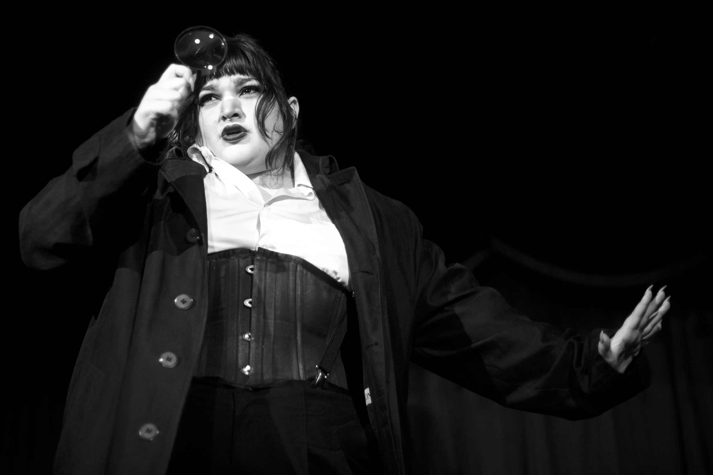 Black and white photo of a woman with dark hair holding a small magnifying glass in front of her, dressed in a dark coat and waist cincher, against a dark background.