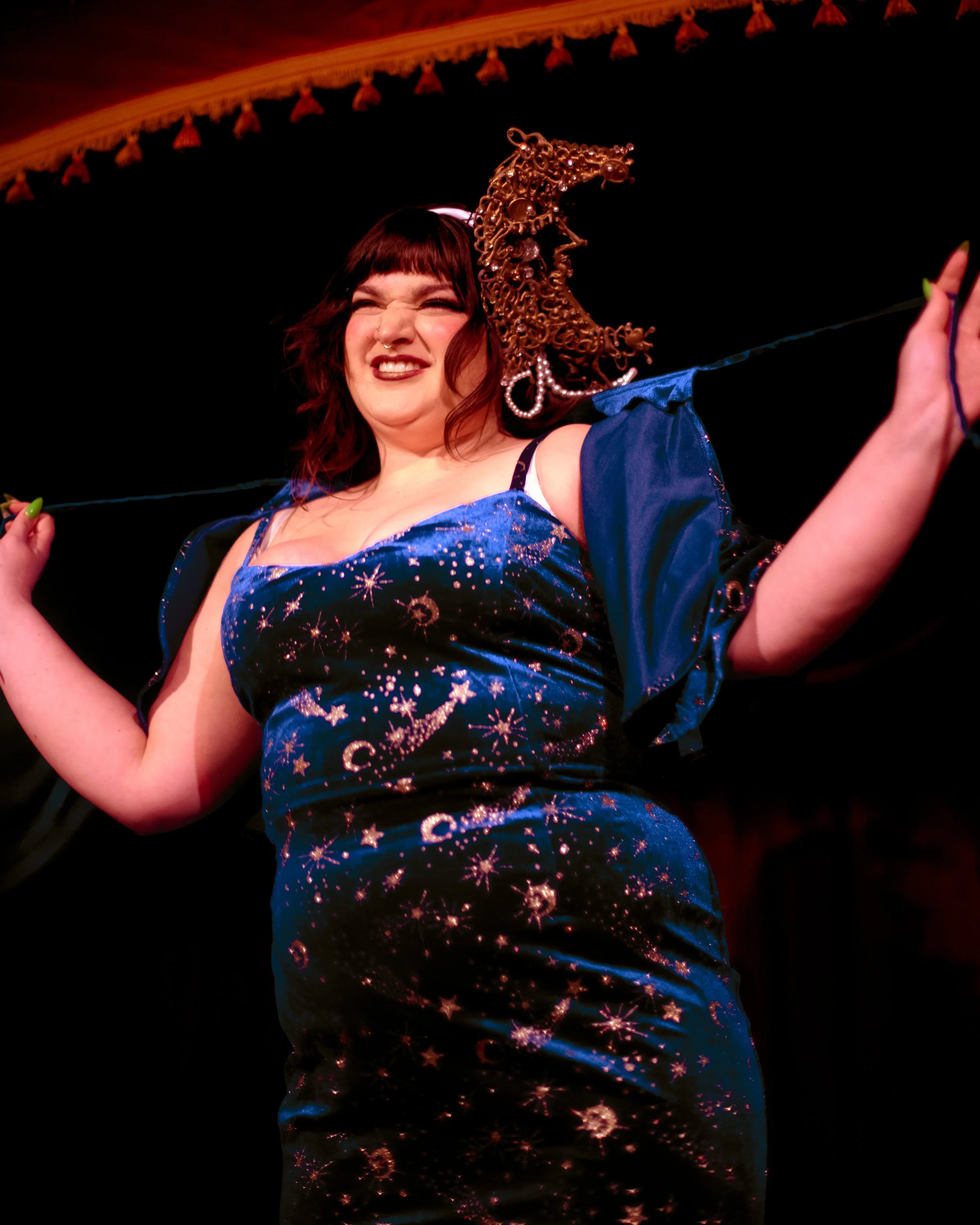 A person smiling and winking, wearing a blue dress with a celestial pattern, with a dark background.