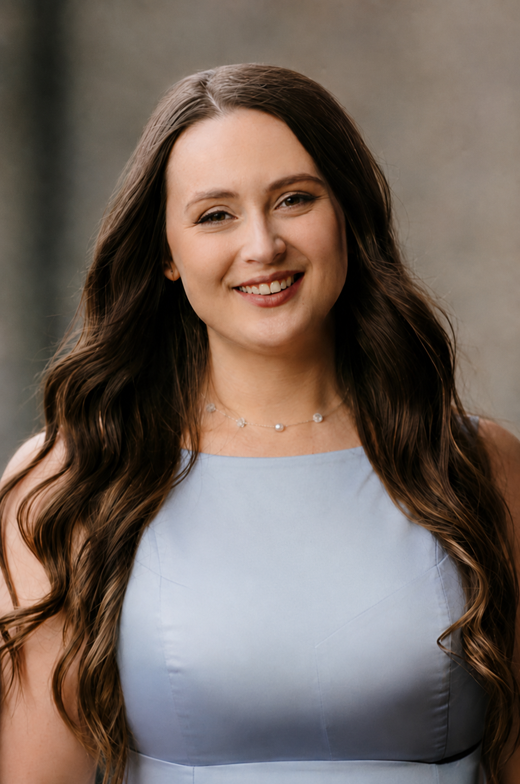 A young woman with long brown wavy hair, wearing a sleeveless light blue dress and a pearl necklace, smiling outdoors with a neutral background.