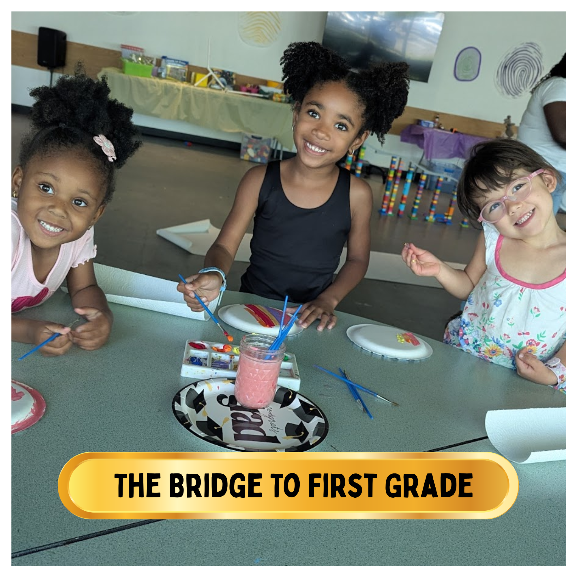 Three young girls smiling while painting at a table in a classroom or activity space, with paintbrushes, paper plates, and art supplies on the table.