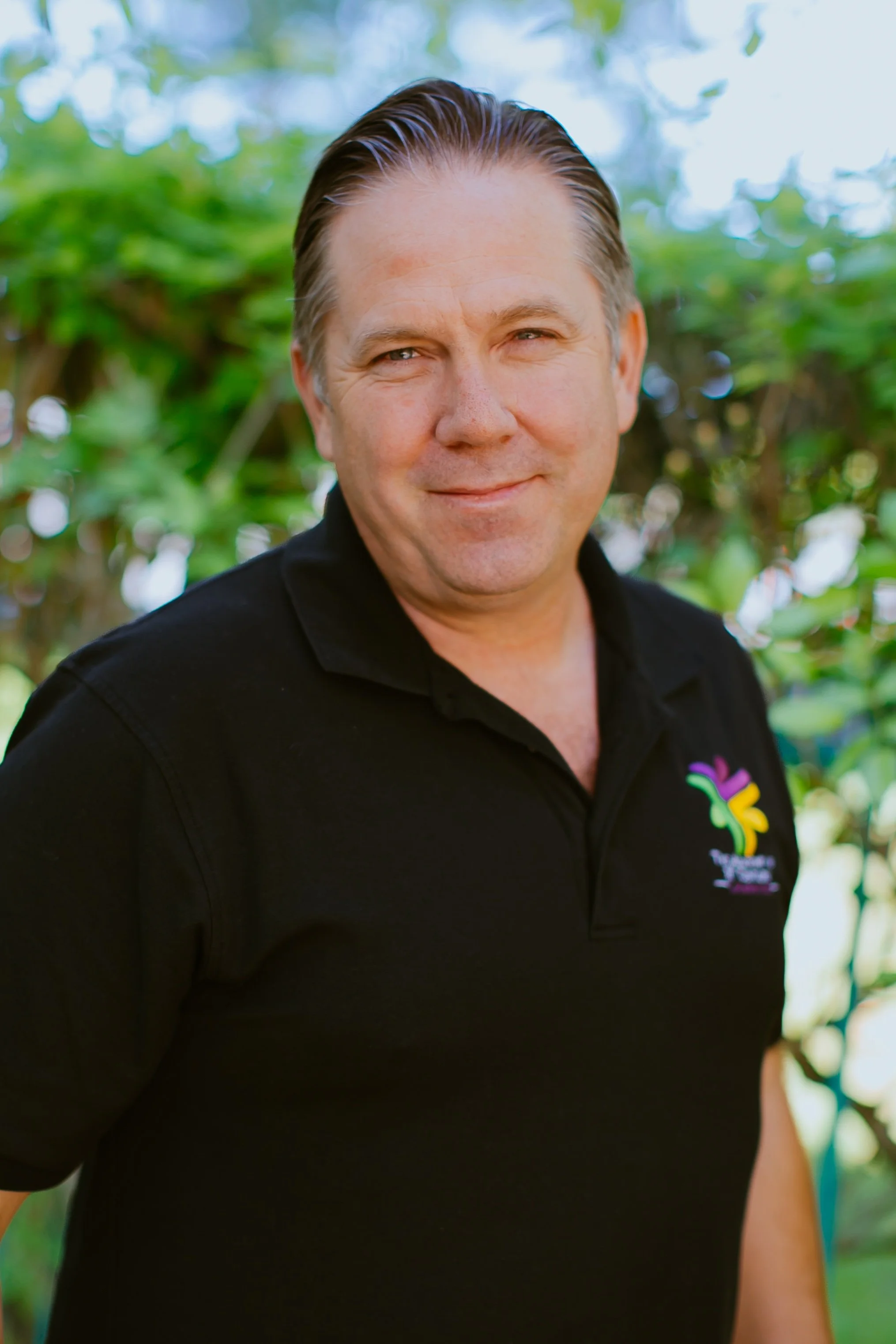 A smiling man wearing a black polo shirt with a colorful logo standing outdoors.