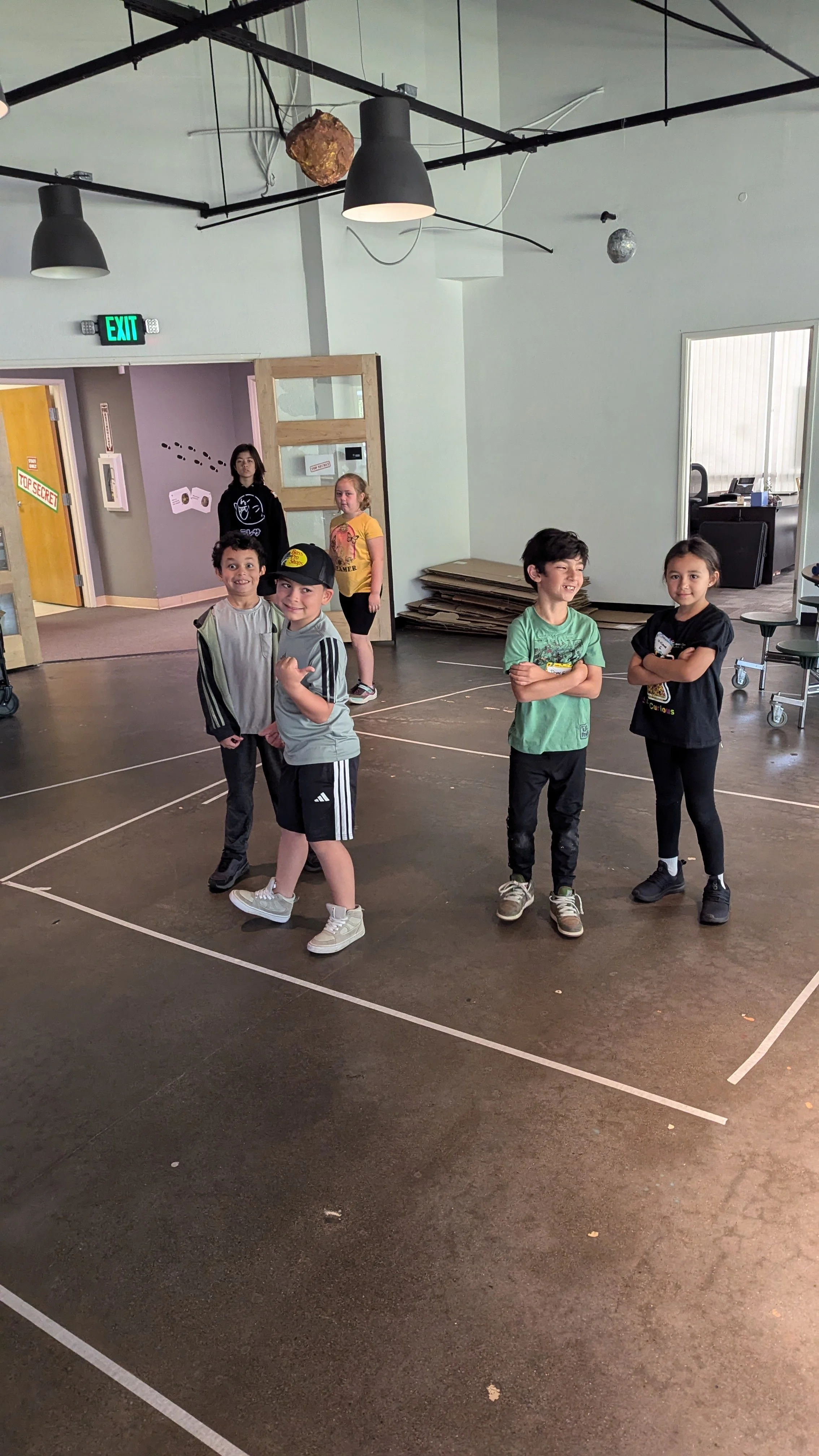 Five children standing on a gym floor in a school or community center, with two boys and three girls, smiling and crossing their arms or making playful gestures. In the background, two more children are standing near the doorway, and there are walls with signs and a stack of cardboard.