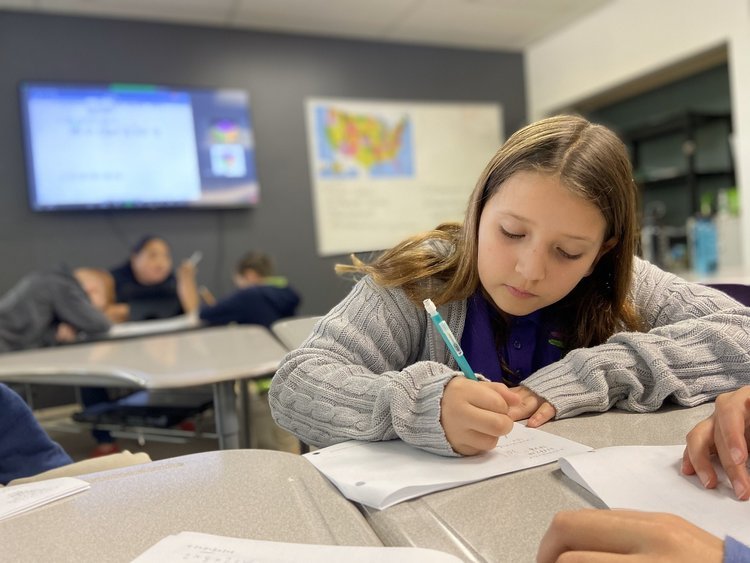 A student in a classroom writing on a paper with a pencil. Other students are visible in the background. The classroom has desks, a wall map, and a digital screen.