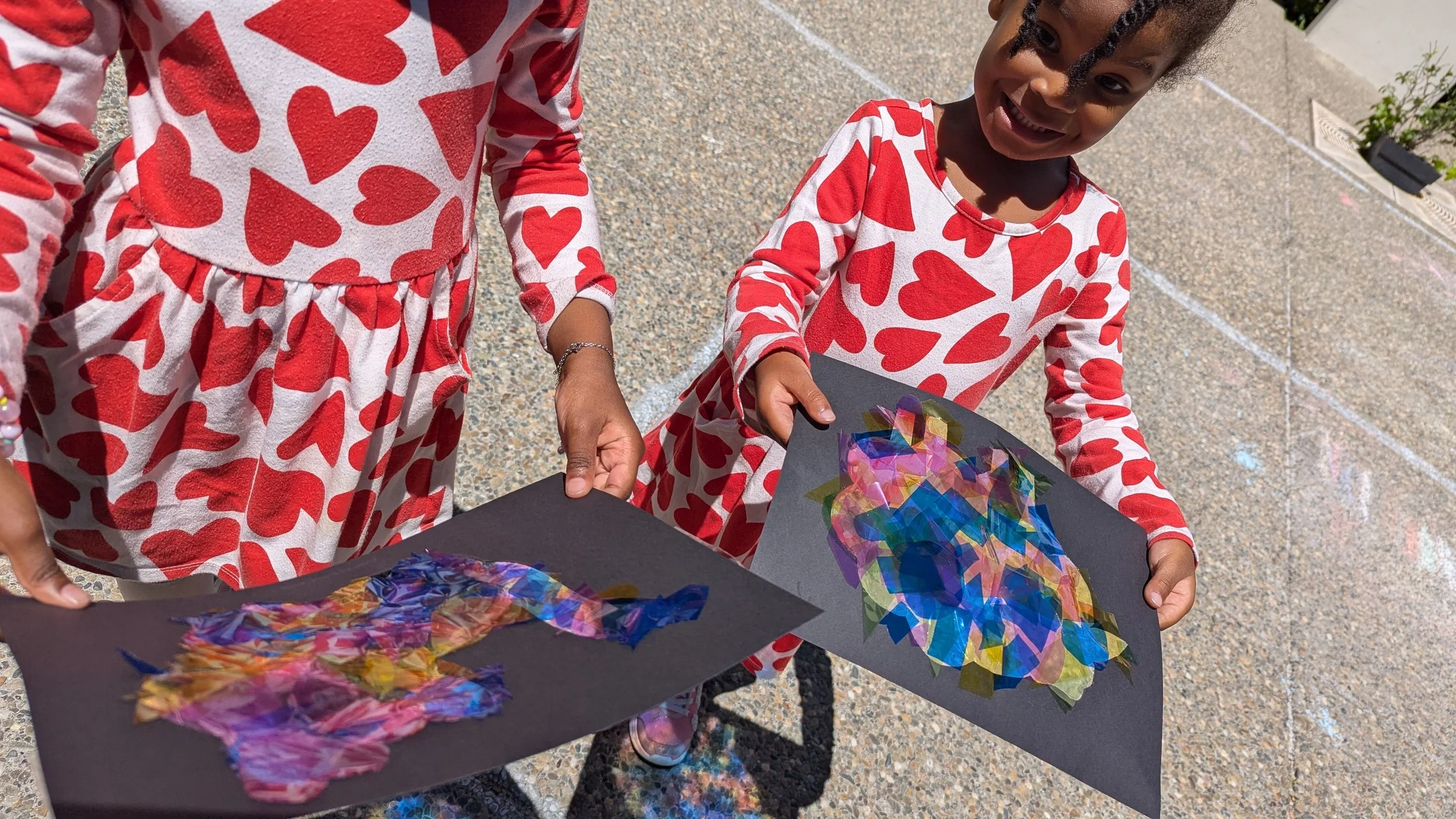 Two young girls in matching red and white heart-patterned dresses holding colorful collaged artwork on black paper, outdoors on a concrete surface.
