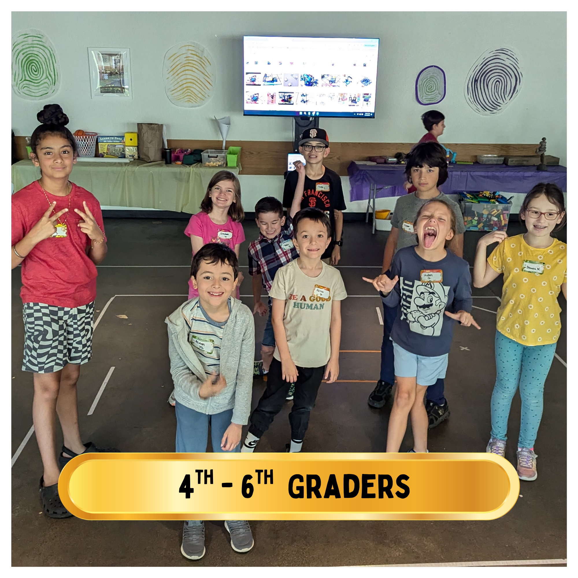 A group of children in grades 4th to 6th posing happily in a classroom or event space with decorations and a large screen in the background. They are smiling and making playful gestures.