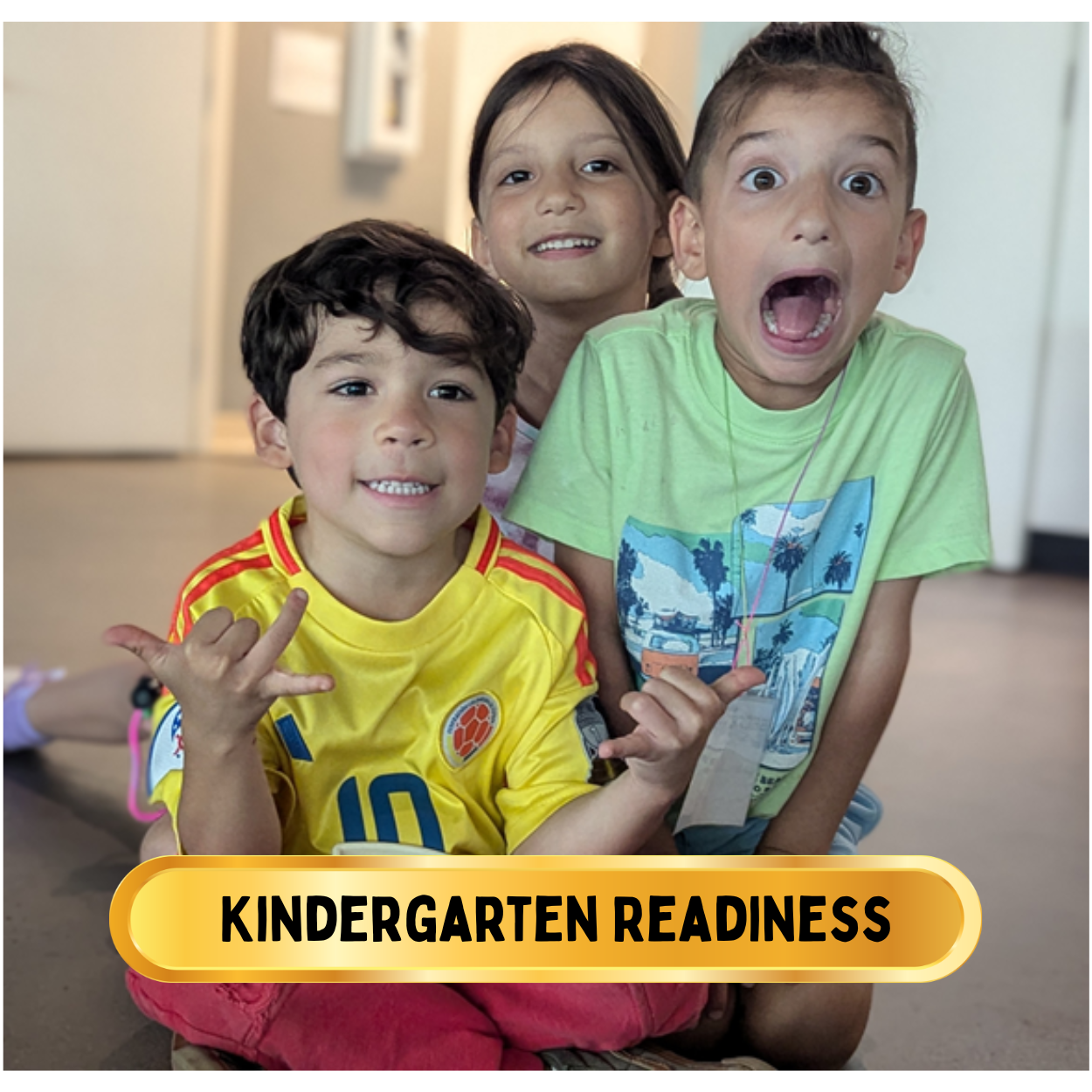 Three young children smiling and making playful gestures, sitting on the floor indoors. The photo is labeled "Kindergarten Readiness."