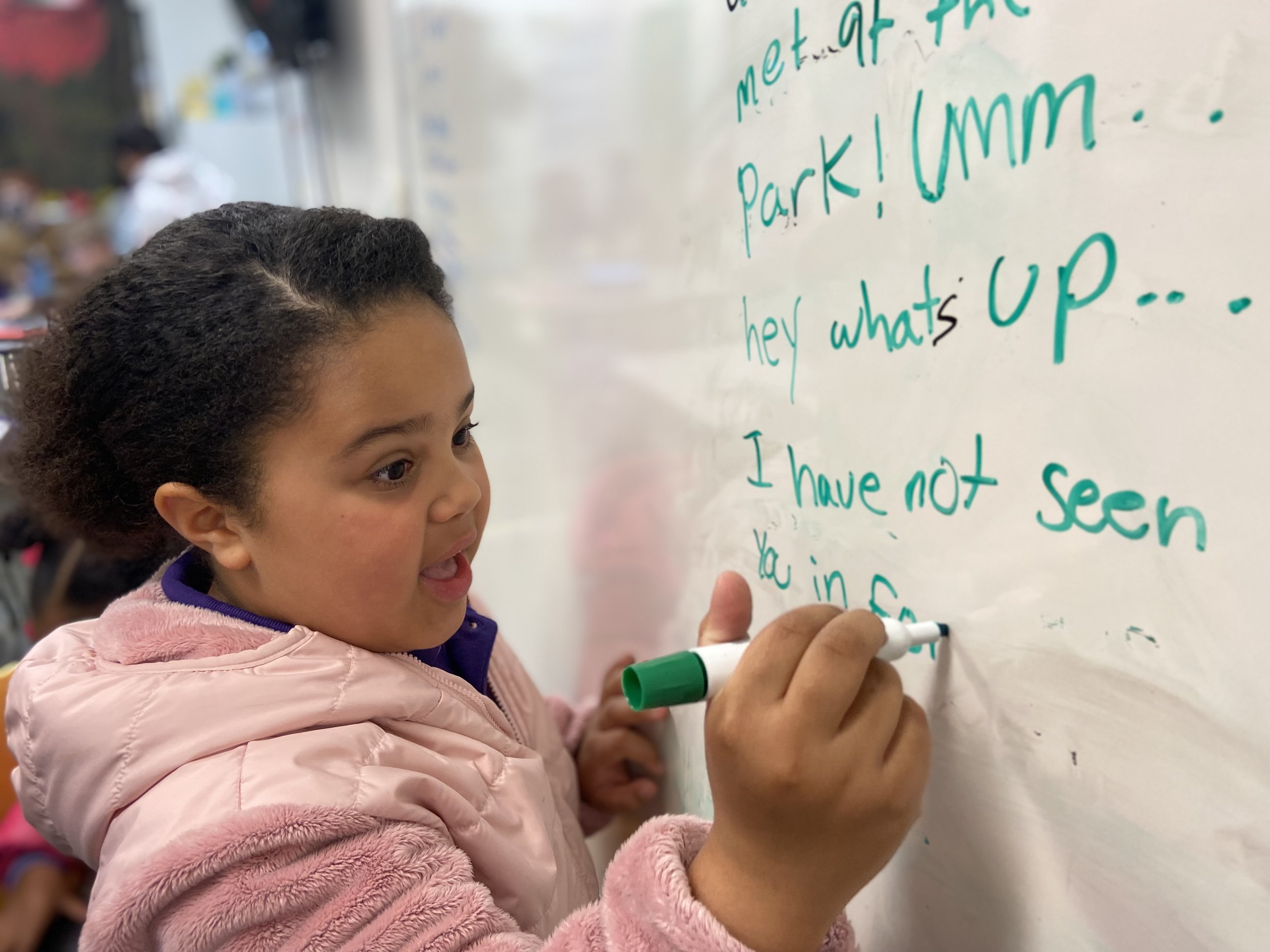 A young girl writing on a whiteboard with a green marker, wearing a pink jacket. The whiteboard has text written in green marker.