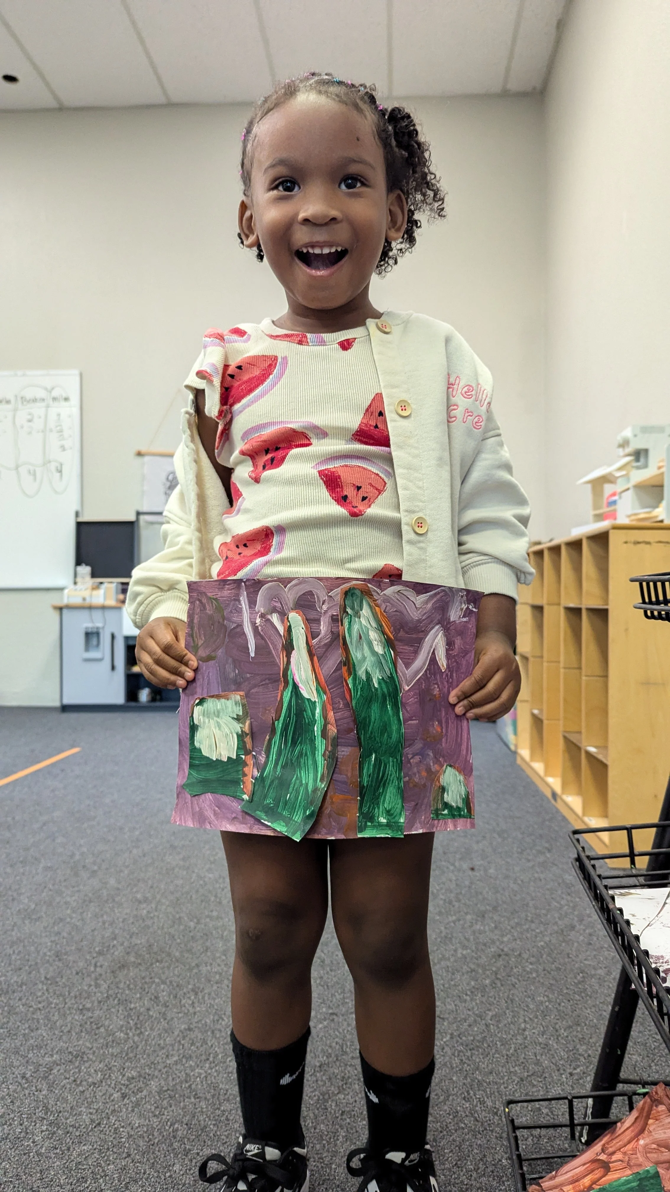 Young girl smiling in a classroom, holding a colorful painted paper art piece.