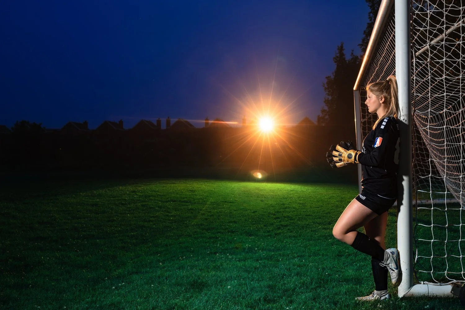 Female soccer goalkeeper standing in the goalpost on a grassy field at dusk, with the sun setting on the horizon.