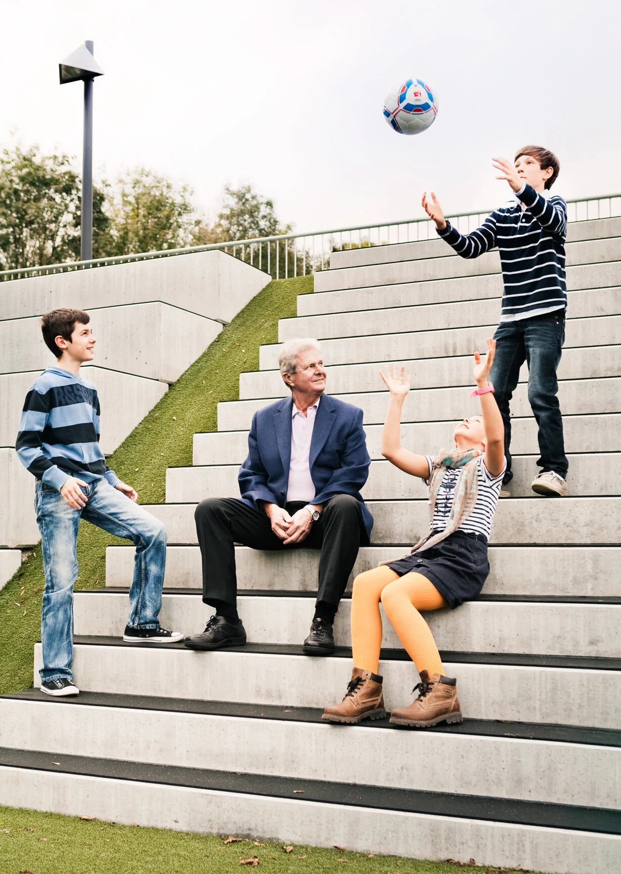 A group of children and an adult man enjoying time outside on concrete stairs, with one boy tossing a soccer ball, and others engaging with him.