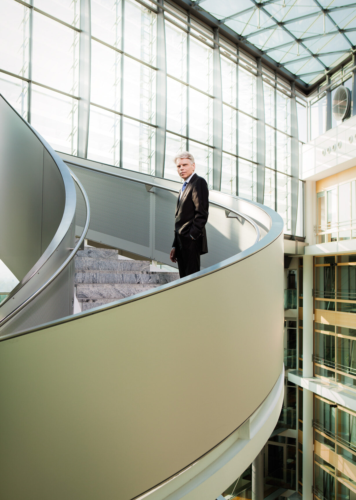 A man in a suit standing on a curved indoor staircase with glass walls and high ceiling in a modern building.