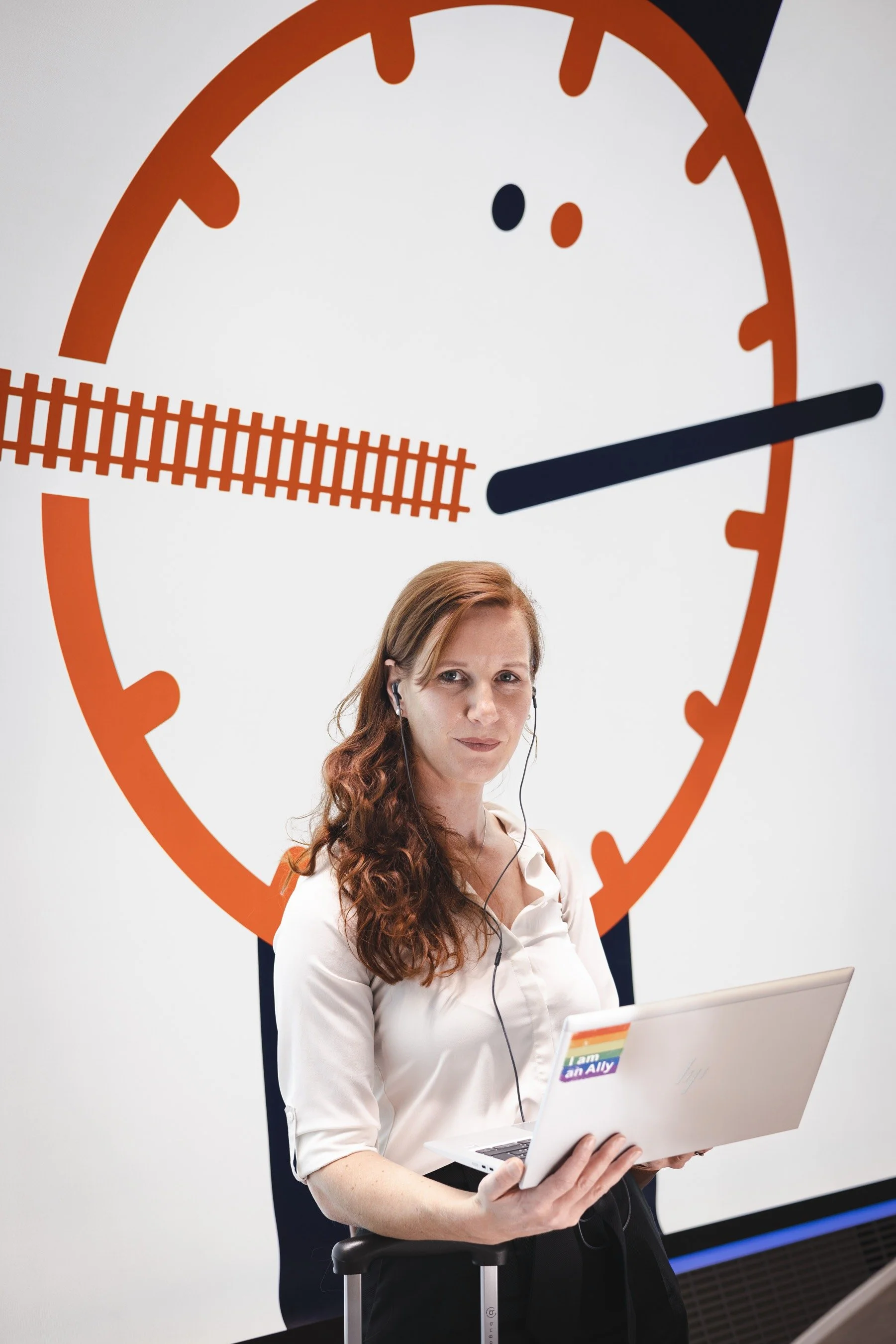 A woman with wavy red hair holding a white laptop with a rainbow pride sticker that says "I am an Ally". She is wearing a white blouse and earbuds. Behind her, there is a large wall clock with orange and black colors, showing the time as approximately 3:15.