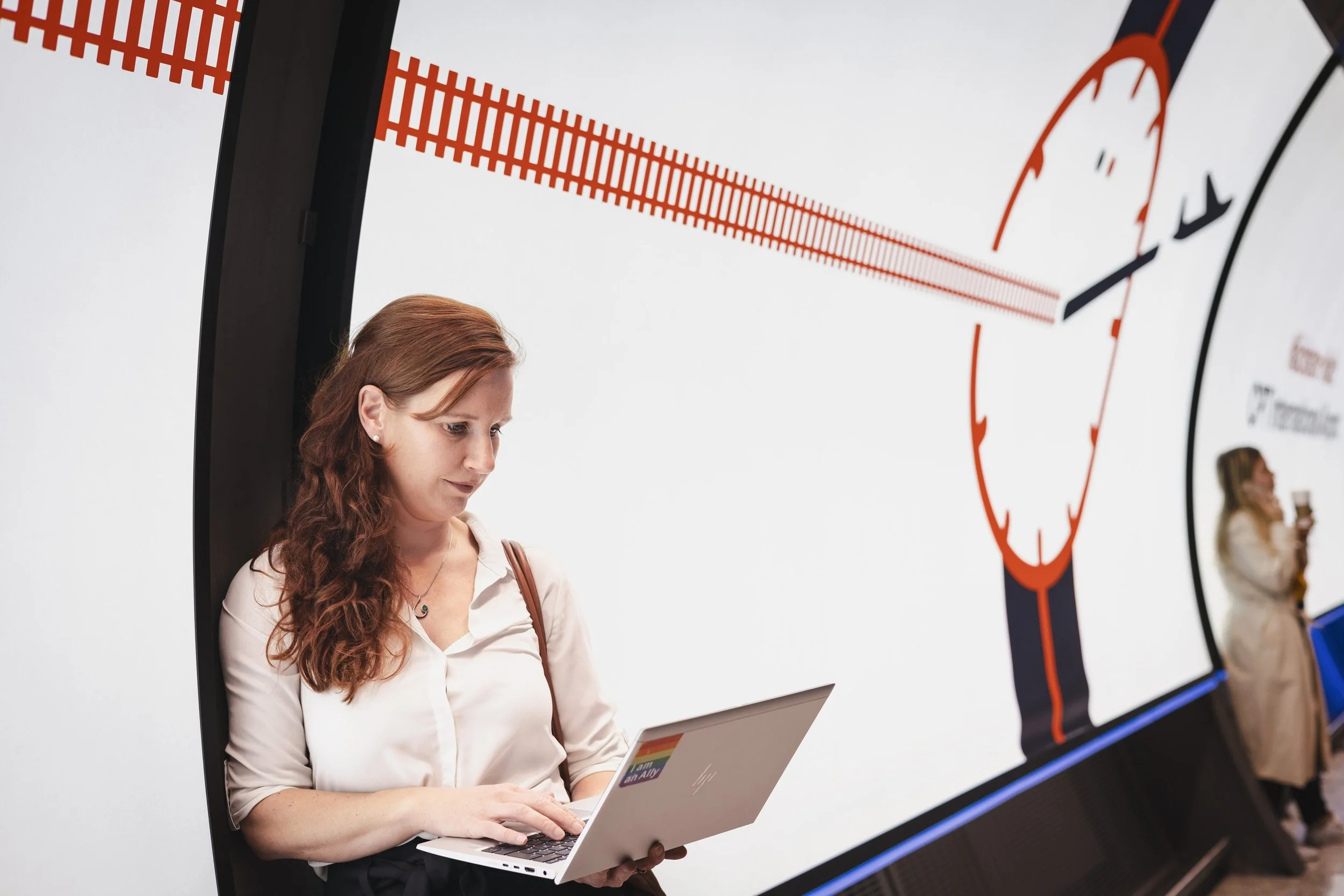 Woman with long red hair using a silver laptop in front of a large digital display showing a clock and a radar with red lines.