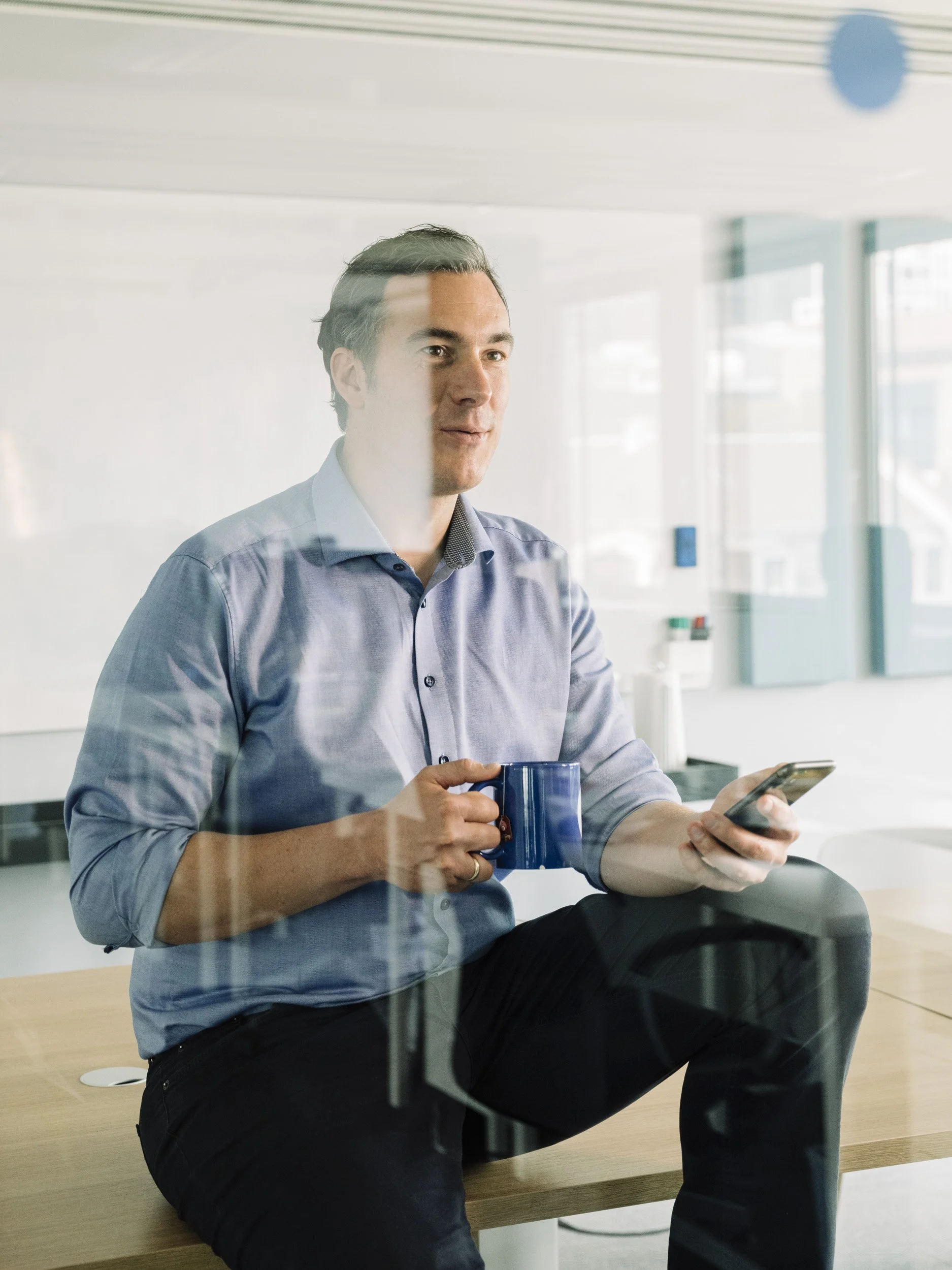 CLARK-Gründer Dr. Christopher Oster sitzt auf einem Tisch und hält ein Handy und eine Tasse in den Händen: Businessfotografie von Angelika Zinzow