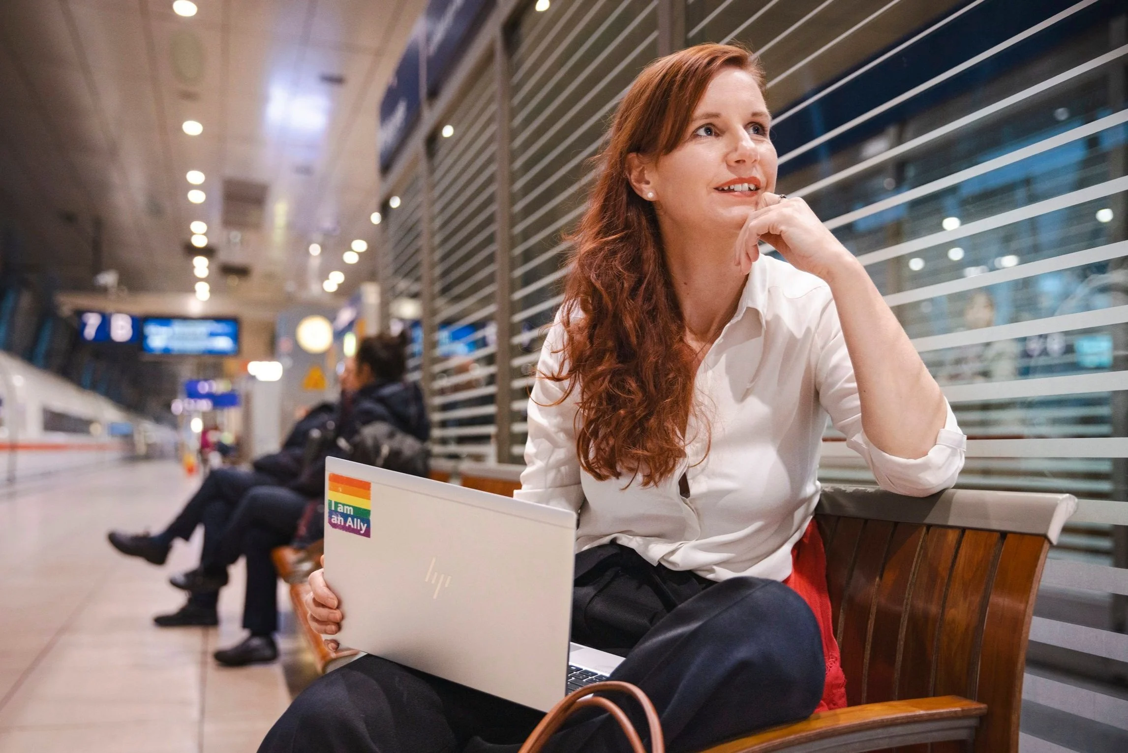 A woman sitting on a bench at an airport, holding a laptop with an rainbow pride sticker that says 'I am an Ally', looking thoughtfully into the distance.