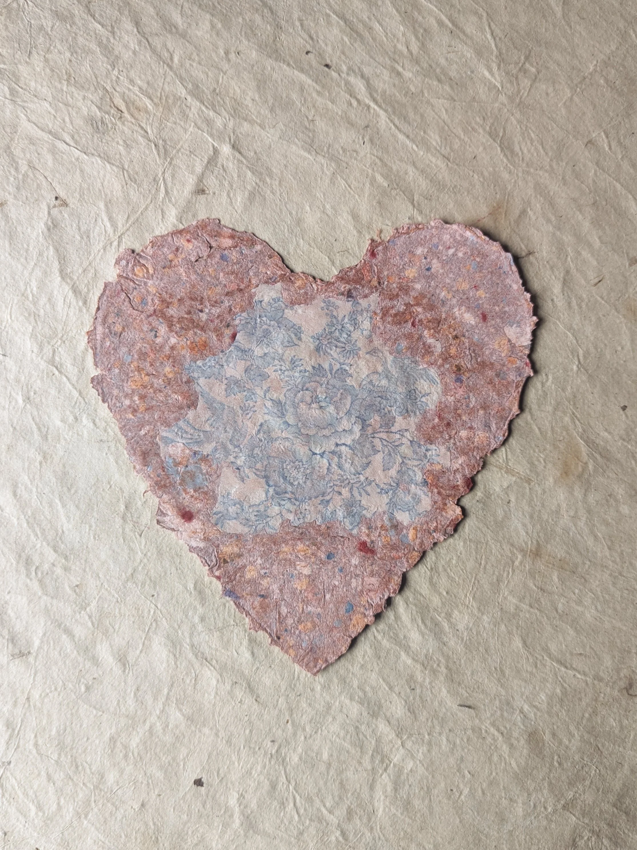 Eine herzförmige Papier  mit einer blauen Pflanzenmuster im Zentrum und einer rostfarbenen, zerknitterten Textur, auf einem beige Papiertisch.