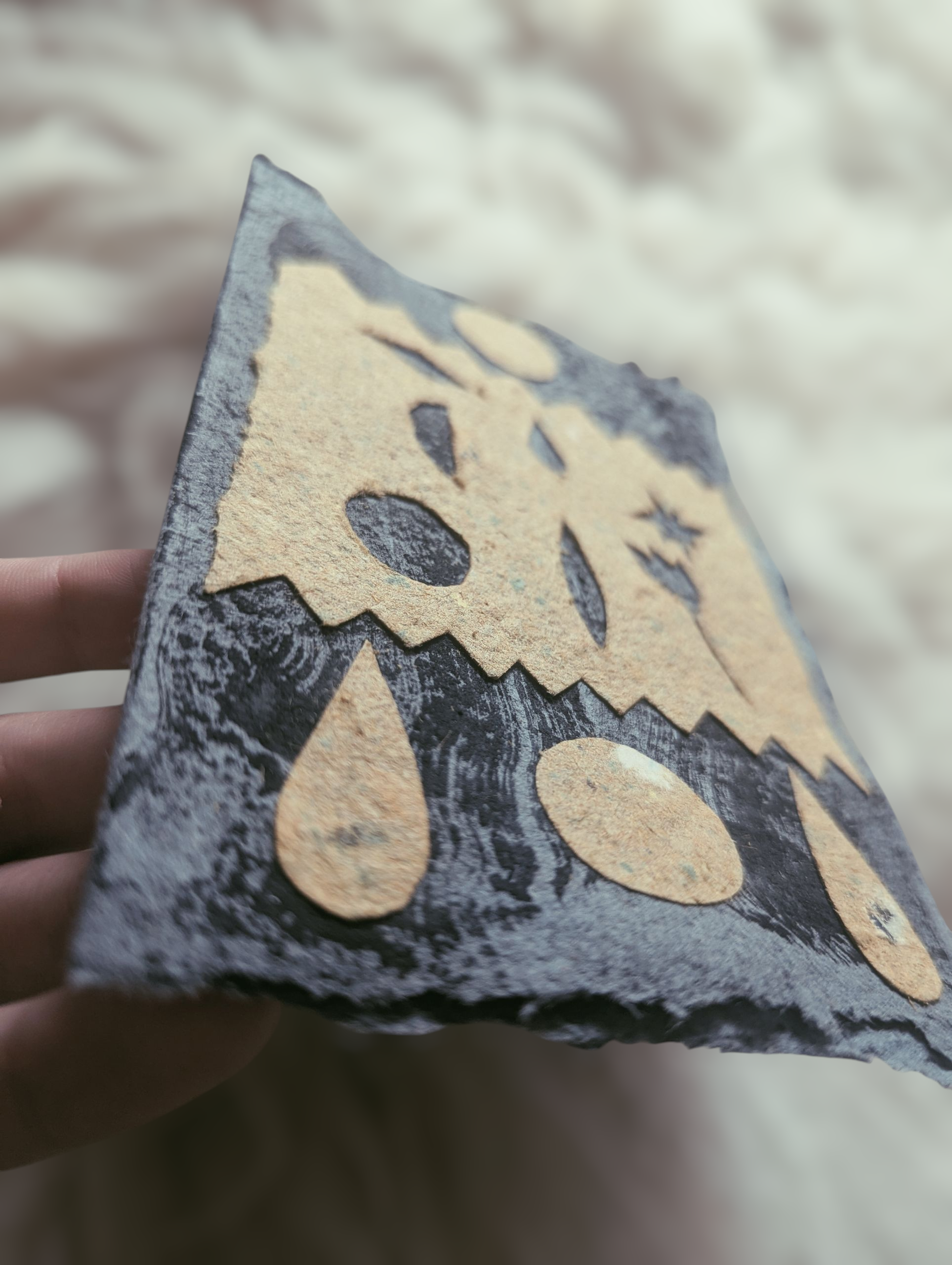 Ein handgehaltes Stück Papier mit einer Halloween-Gesichtsschablone, das wie ein Kürbis mit Tropfen und ovalen Formen aussieht.