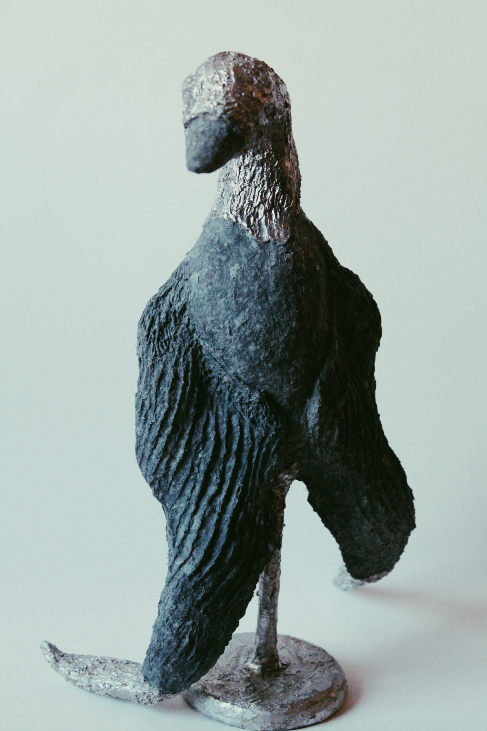 Eine Bronzefigur eines stehenden Vogels mit langer Hals und Schnabel, auf einem runden Sockel. Die Oberfläche ist texturiert und weistDetails auf.