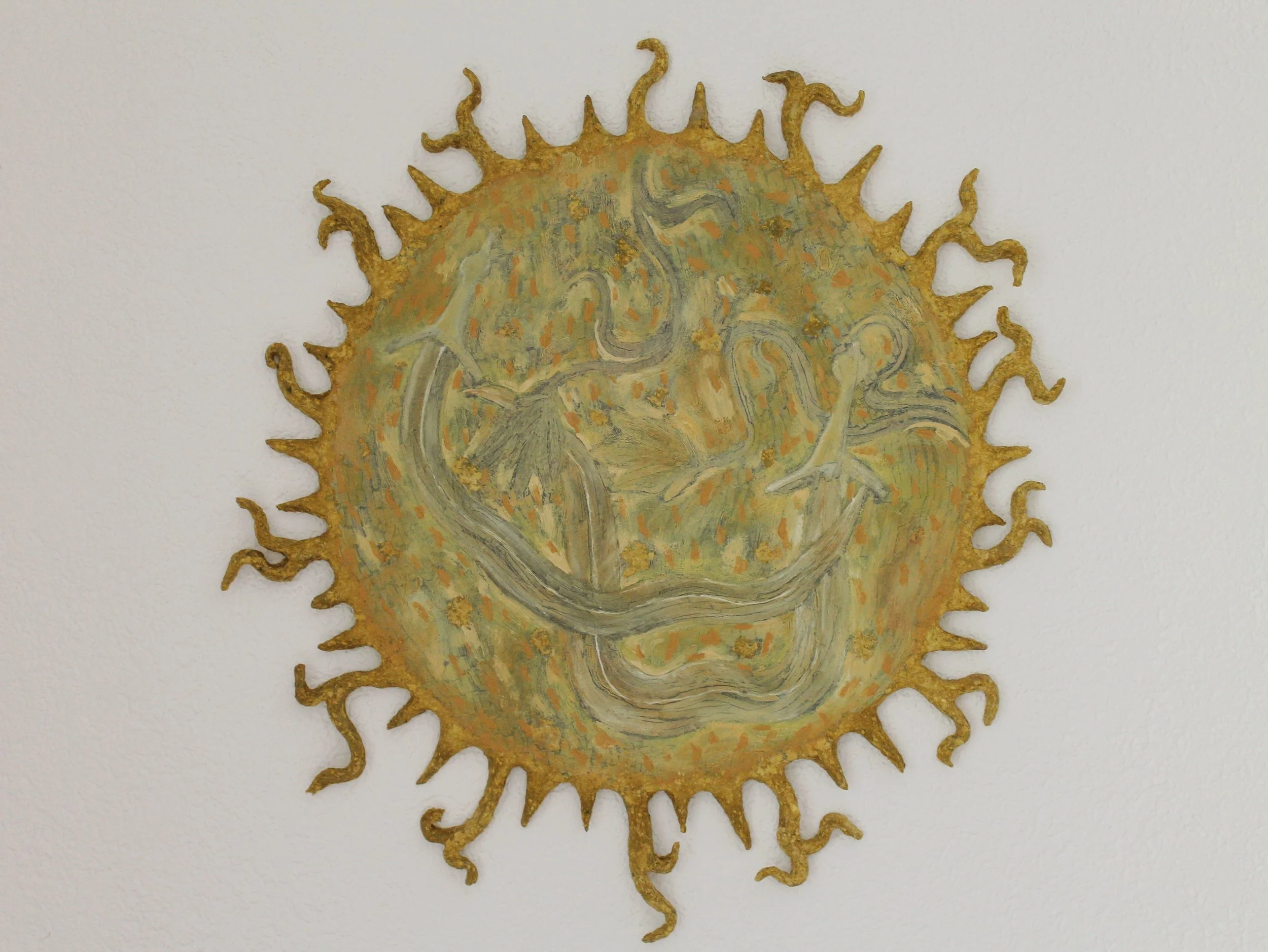 Metalische Kunstinstallation in runder Form mit stilisiertem Tiergesicht und welligen, tentakelartigen Anhängen an der Außenseite, vor weißer Wand.