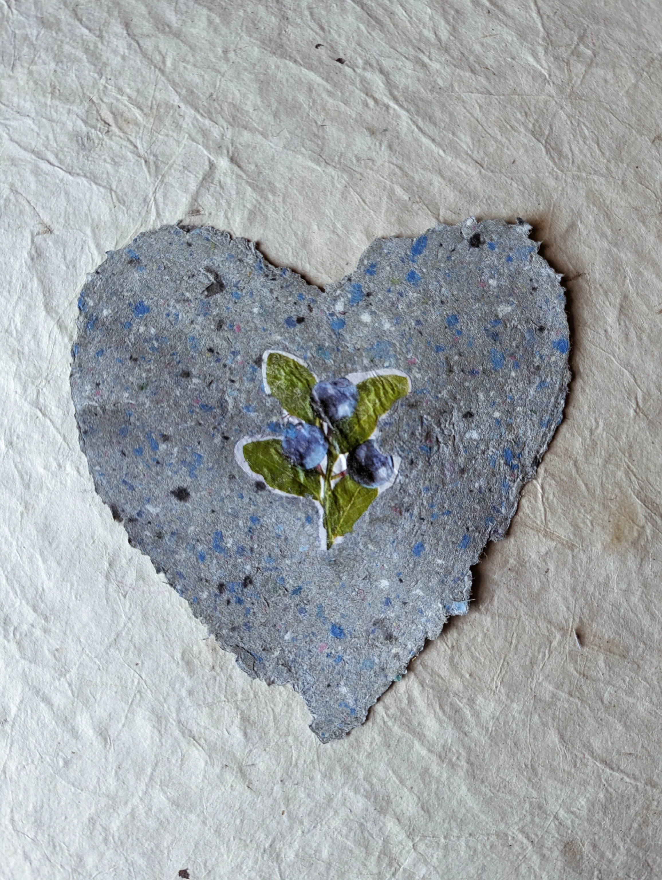Herzförmiges handgeschöpftes Papier  mit kleinen blauen Beeren und grünen Blättern in der Mitte, auf einer Papierunterlage