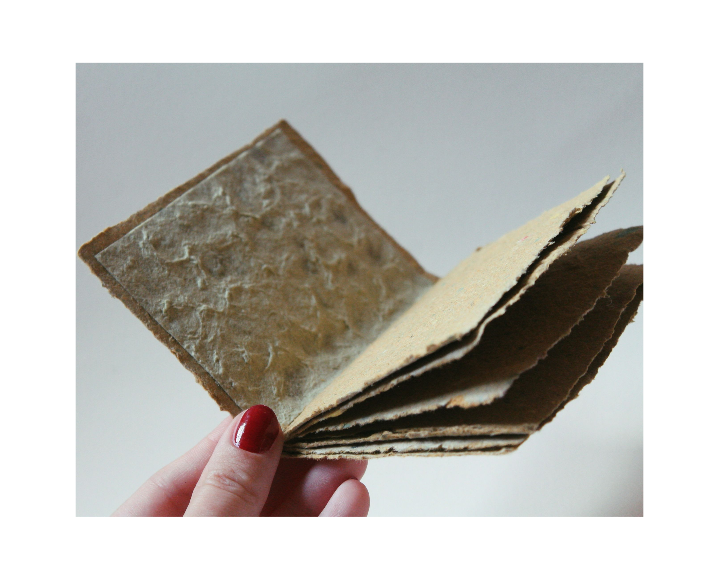 Hand mit roten Nägeln hält ein gefaltetes Buch aus Naturpapier gegen eine weiße Wand.