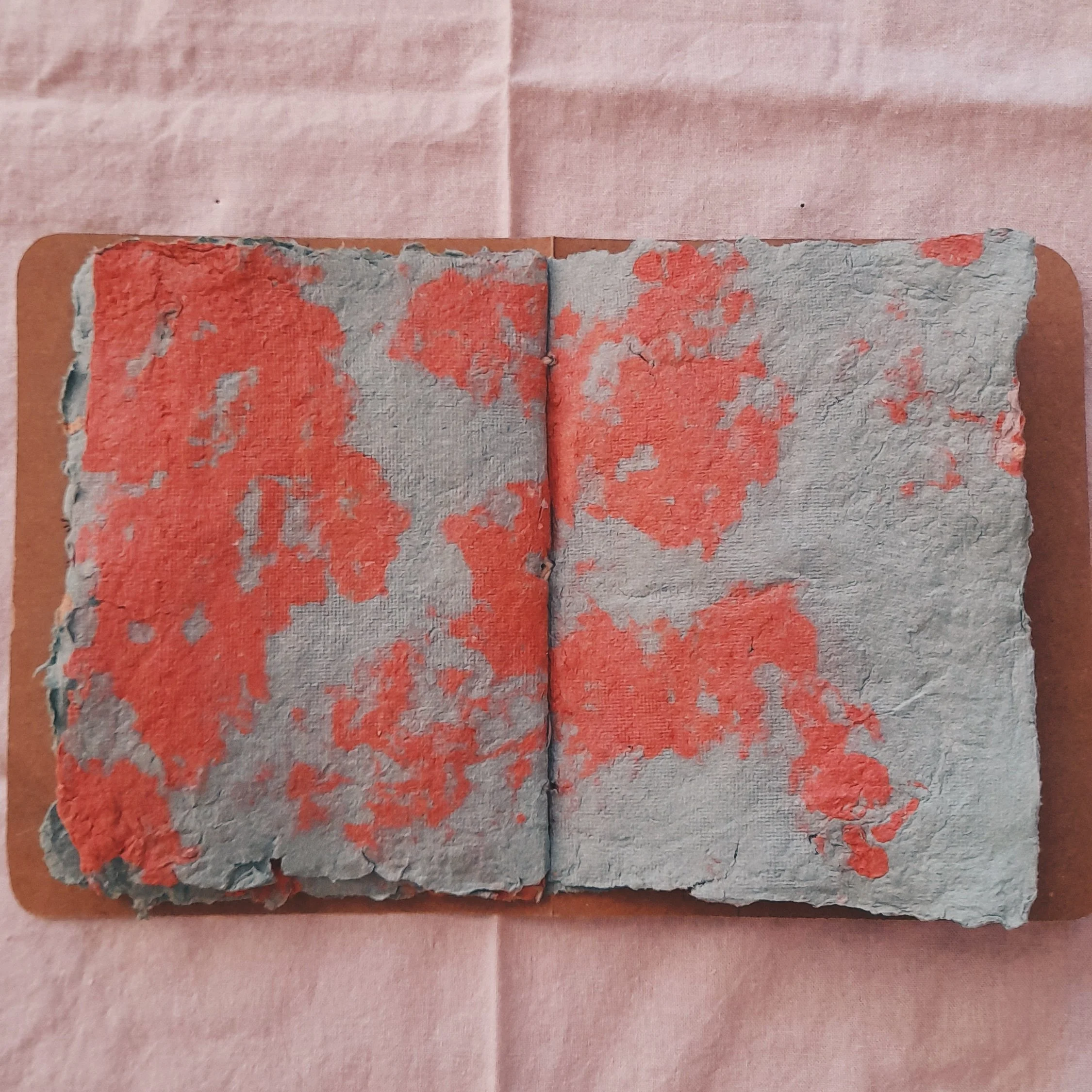Alte, zerfetzte Papierseiten mit orangefarbenen Flecken, auf braunem Untergrund und rosa Tischdecke.