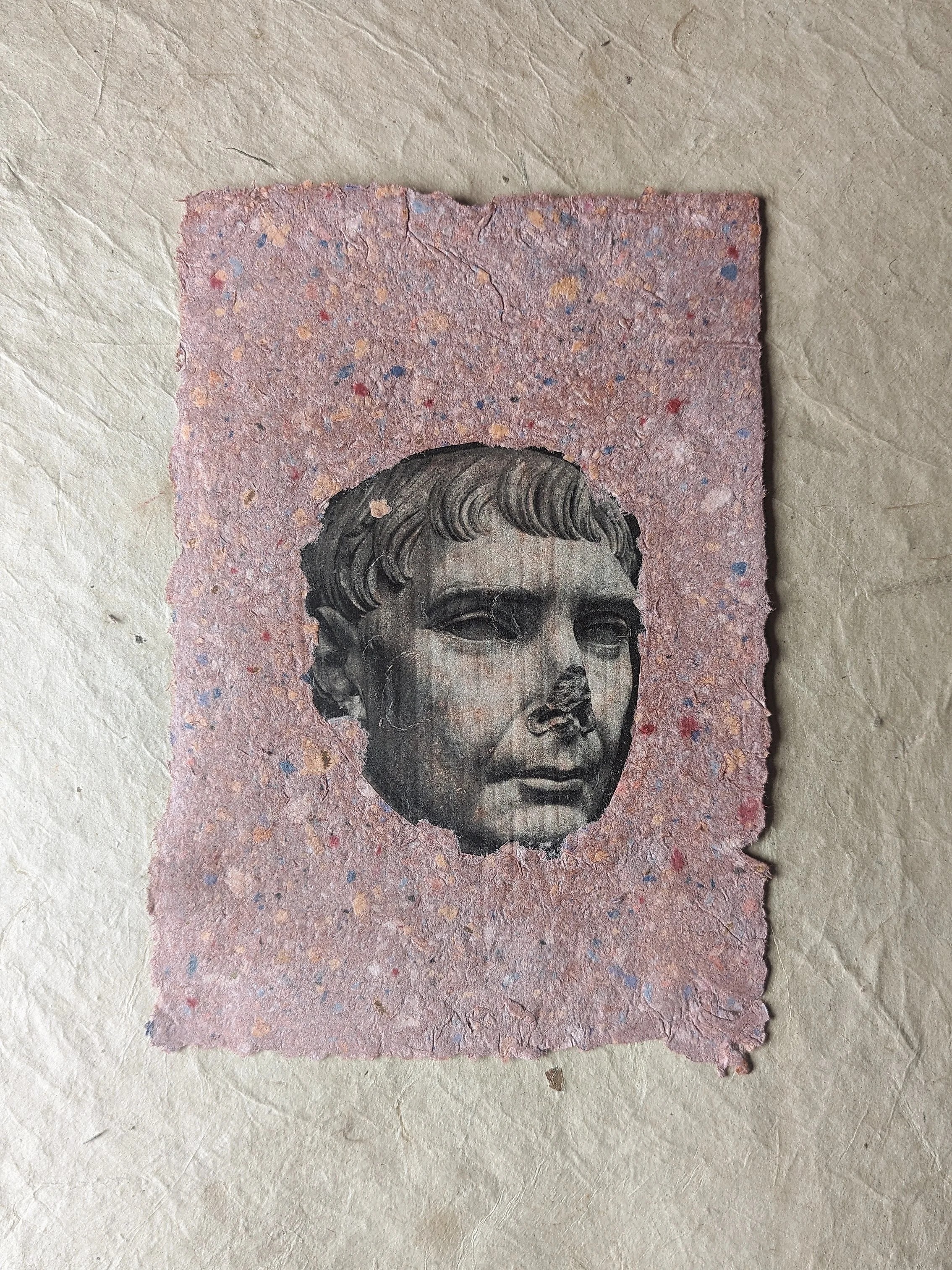 Eine kleine, rechteckige Papierkarte mit einem schwarz-weißen Portrait eines jungen Mannes in der Mitte, umgeben von bunter, getrockneter Farbe auf rosa Hintergrund.