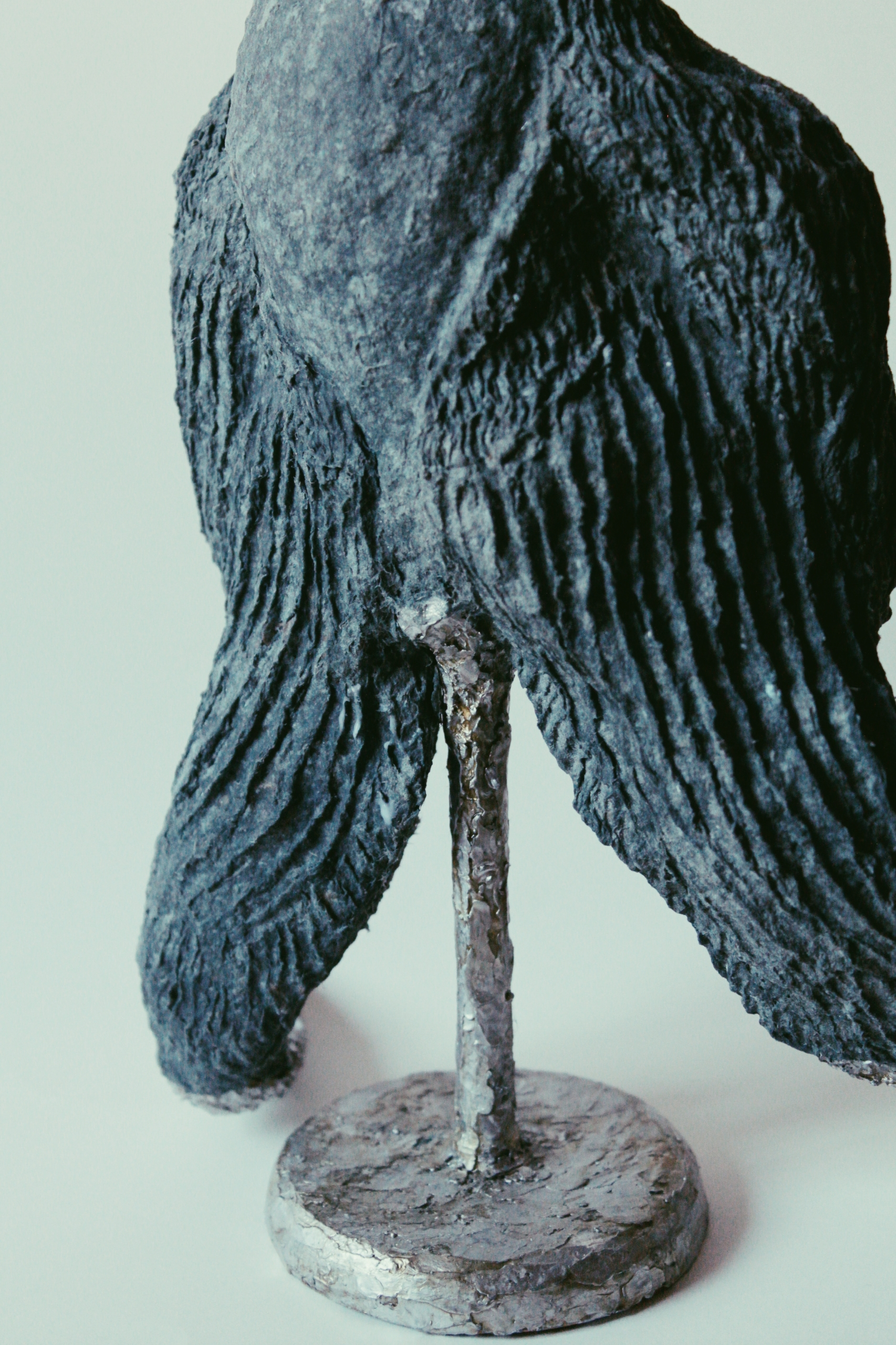 Skulptur eines Vogels mit detaillierten schwarzen und grauen Federn, auf einem runden, grauen Sockel, gegen einen hellen Hintergrund.