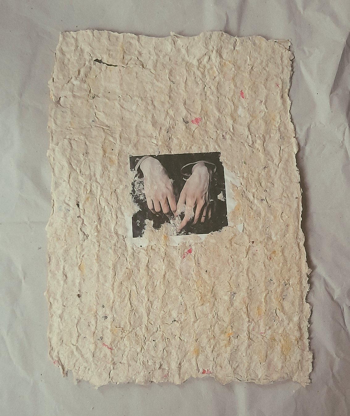 Ein  handgeschöpftes Papier mit einem schwarzen und weißen Bild eines Hands, die eine kleine Figur oder eine ähnliche Form berühren, im Zentrum.