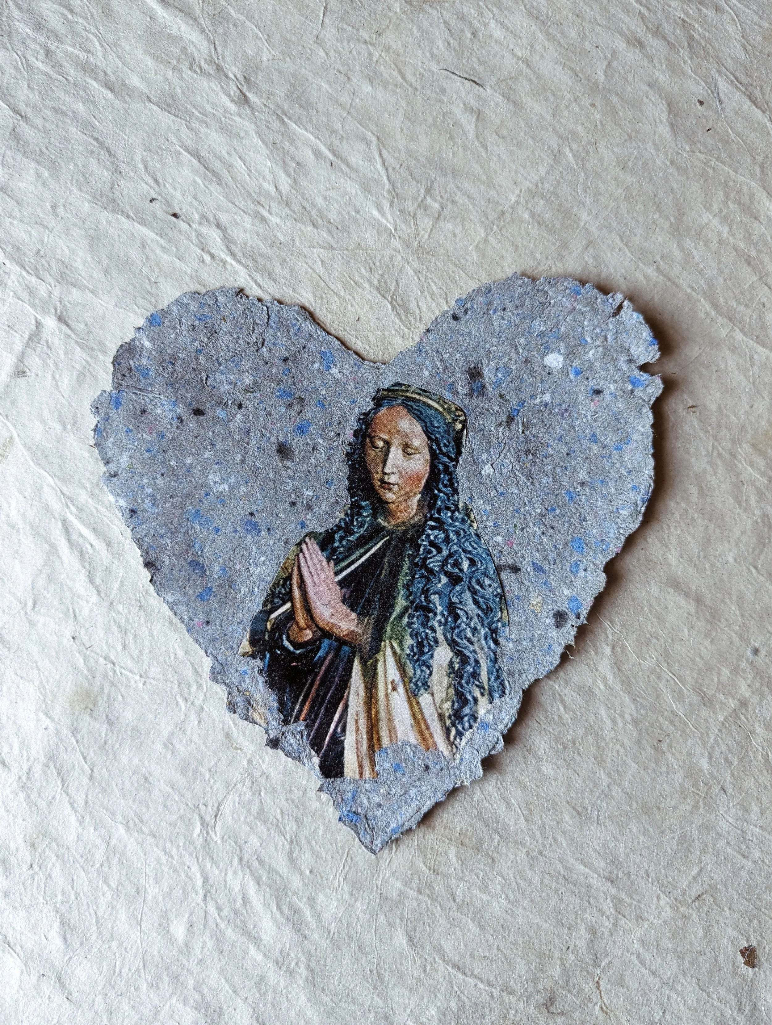 Ein Herz aus Papier mit einem religiösen Bild einer Frau mit verschränkten Händen, das aussieht wie eine Heilige, auf der Oberfläche.