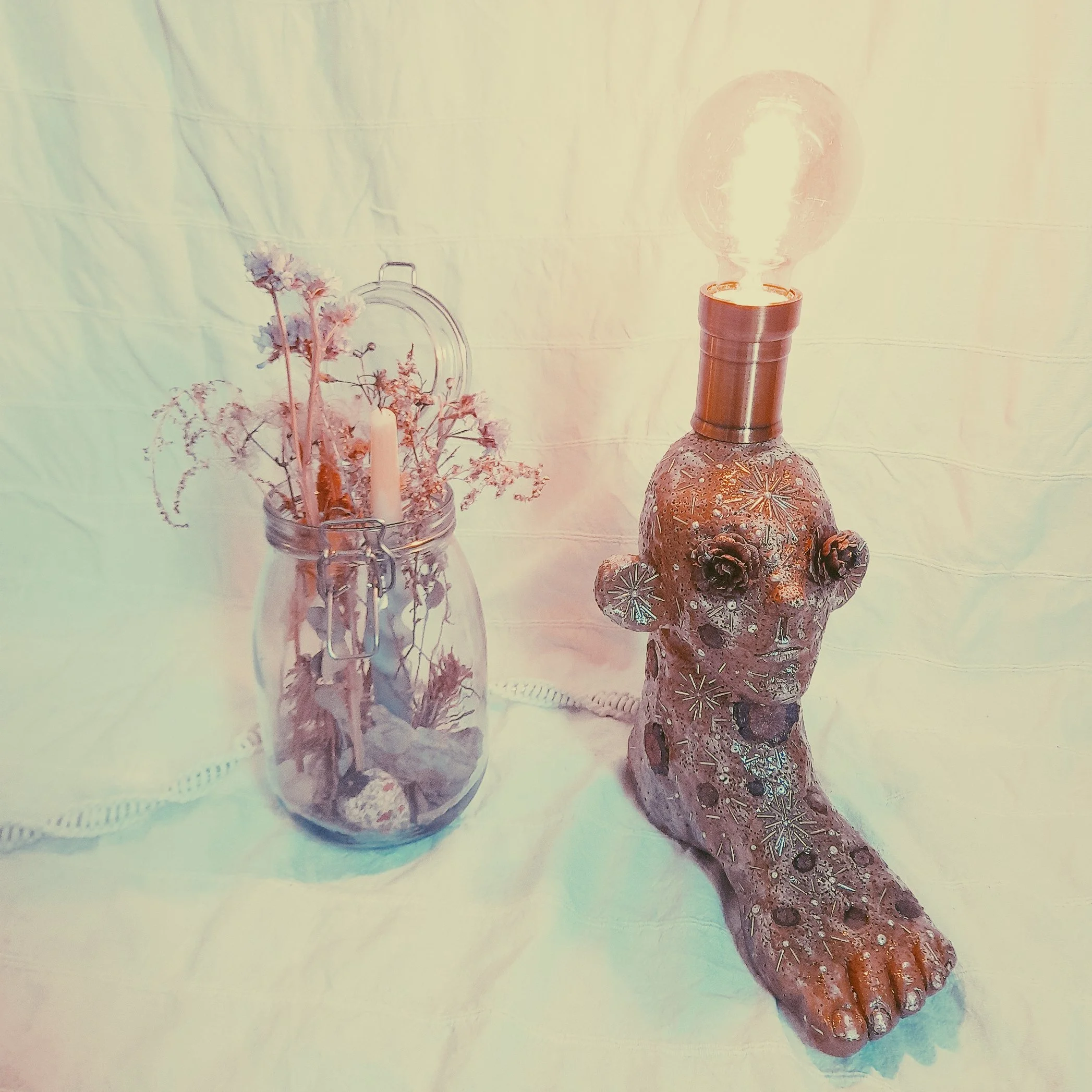 Ein dekorativer Tisch mit einer Glaskaraffe mit Blumen, einer dekorativen Vase mit getrockneten Blumen, einem Kerzenleuchter, einem Kerzen und einer großen, mit Mustern verzierten Vase, die möglicherweise als Lampe dient.